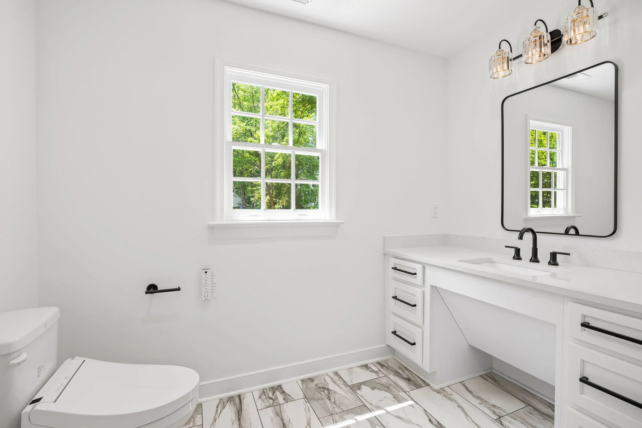 Bright bathroom with white walls, marble floor tiles, a window showing green trees outside, a white vanity with black handles, a large black-framed mirror, modern black faucet, a white toilet, and wall-mounted lighting fixtures.