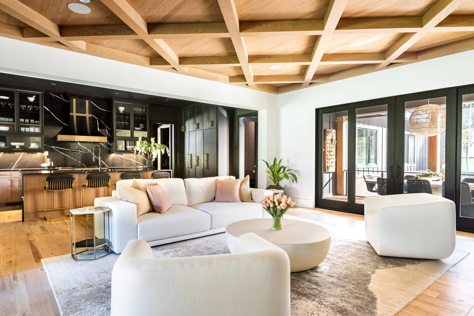 Modern living room with white sofa, armchair, round coffee table with flowers, black kitchen cabinets with marble backsplash, sliding glass doors leading to a patio.