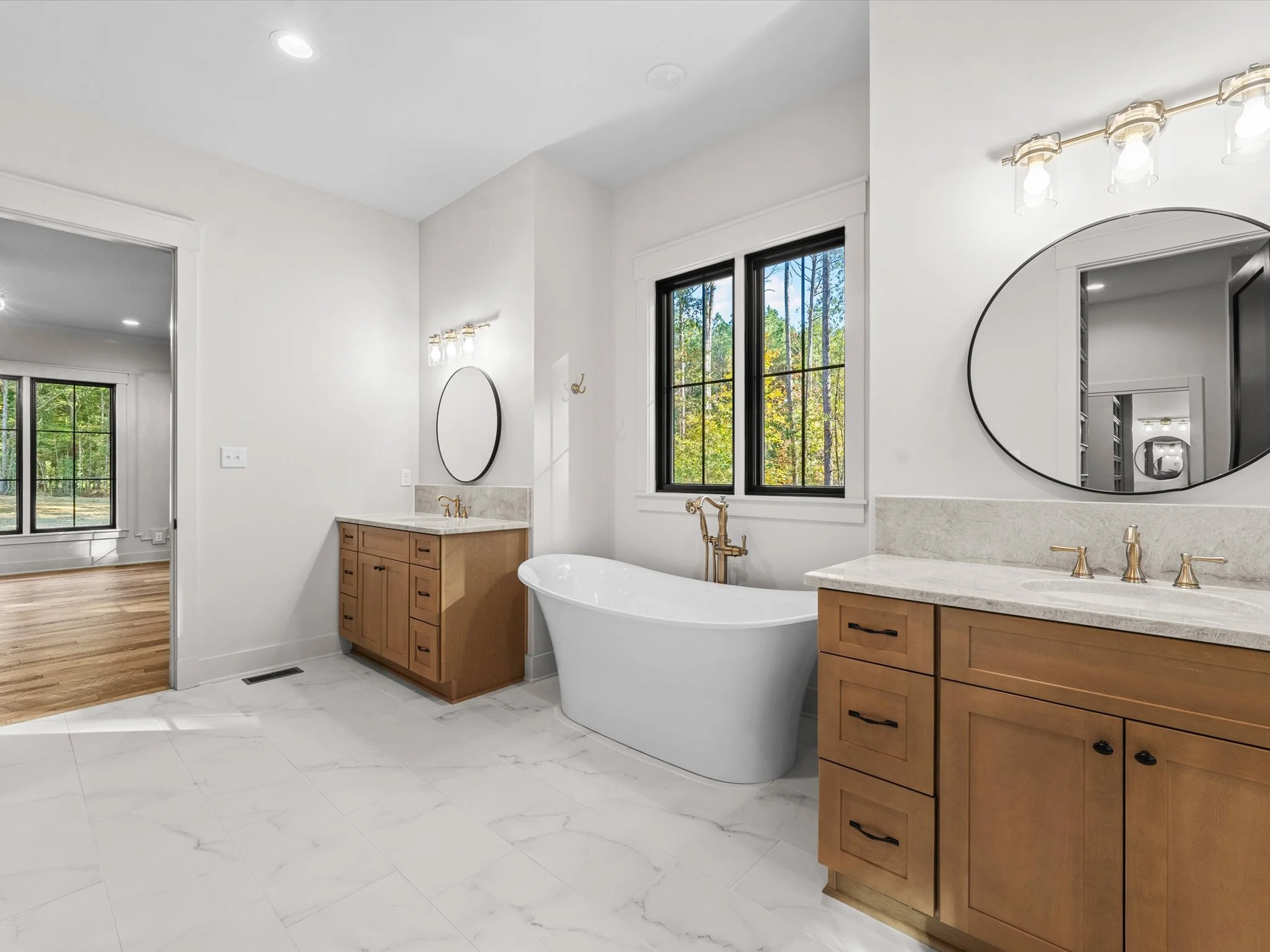 Modern bathroom with two wooden vanities, a freestanding bathtub by the window, and two oval mirrors above the vanities, with black-framed windows showing an outdoor view.