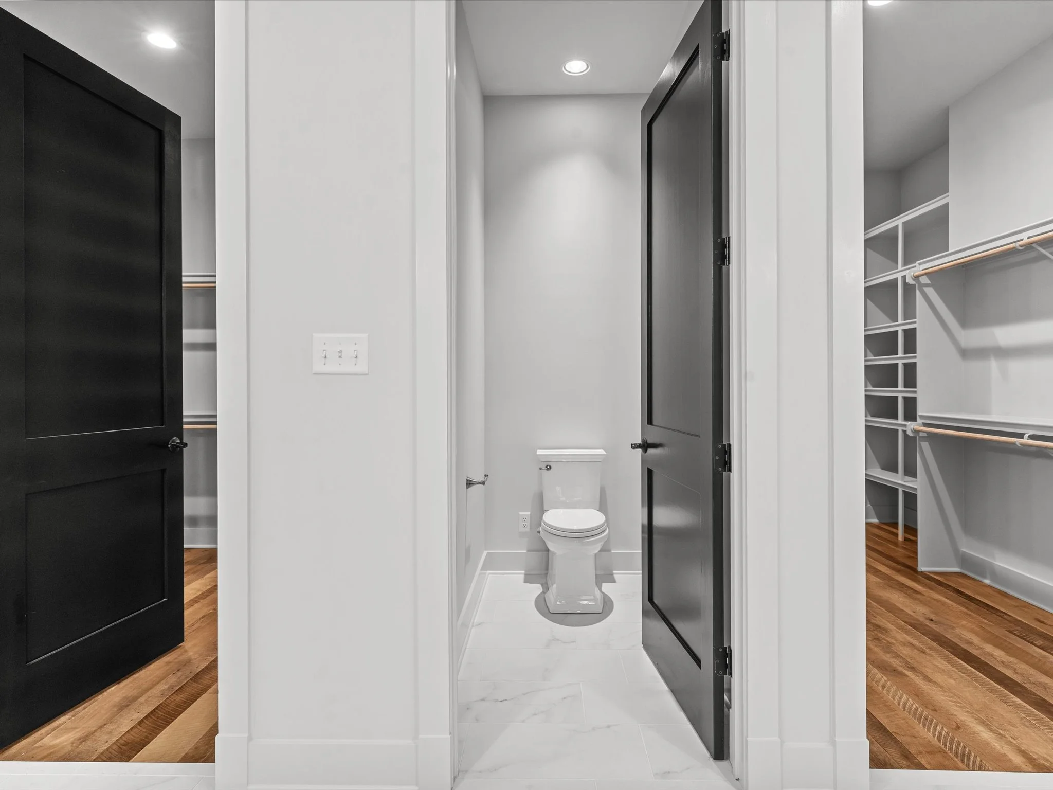 A modern bathroom with a white toilet, black doors, and a walk-in closet with wooden shelves and hanging rods.