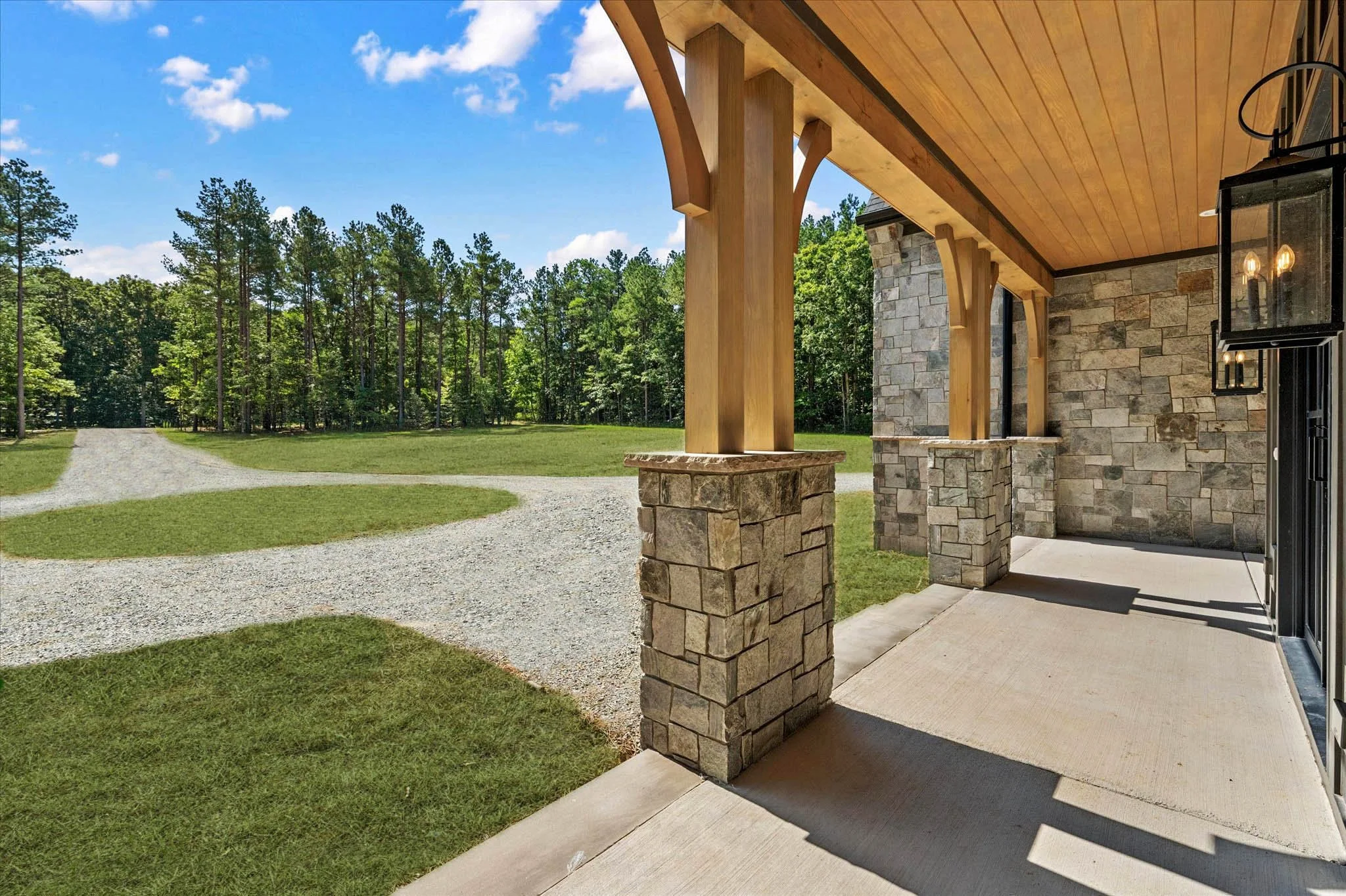 A front porch of a house with stone and wood construction, outdoor lanterns, and a gravel driveway leading to a green lawn and a wooded area under a partly cloudy blue sky.