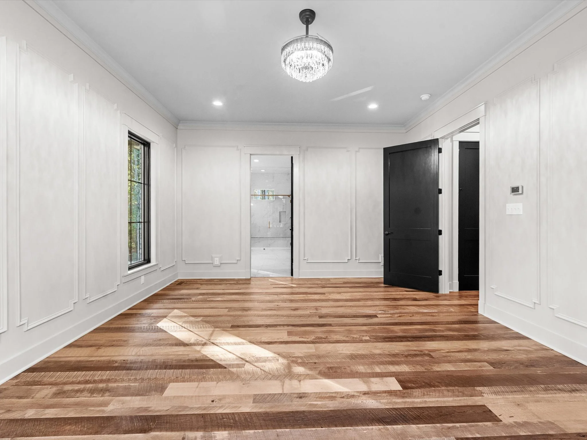 Empty room with white paneled walls, hardwood flooring, large windows, black door, and ceiling light
