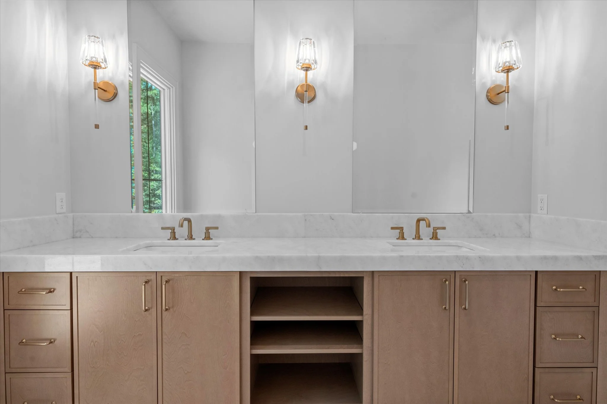 Bathroom vanity with a large mirror, two gold faucets, and a marble countertop, flanked by two gold wall sconces with glass shades.