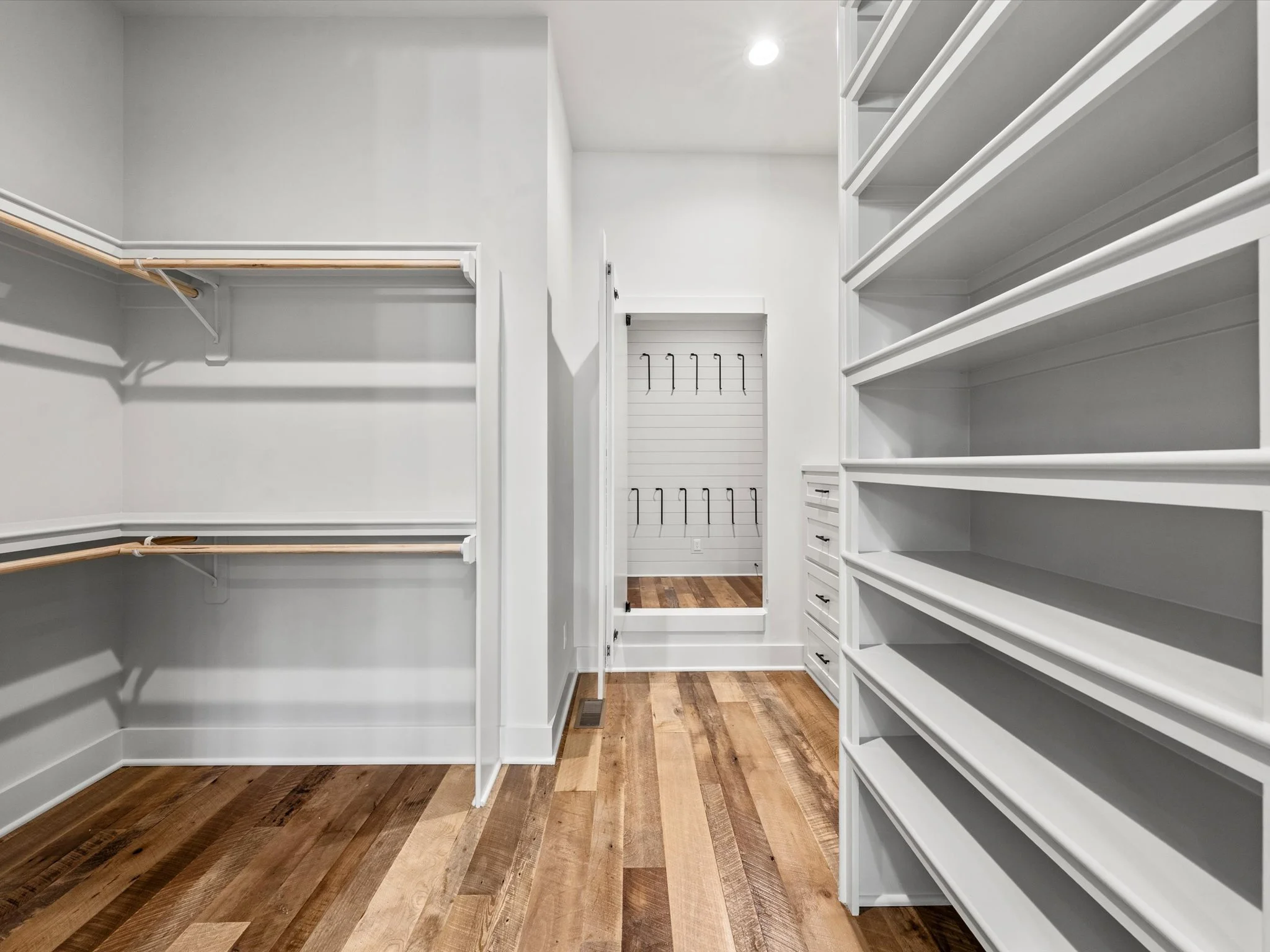 Empty walk-in closet with white built-in shelves and drawers, hardwood floors, and space for hanging clothes.