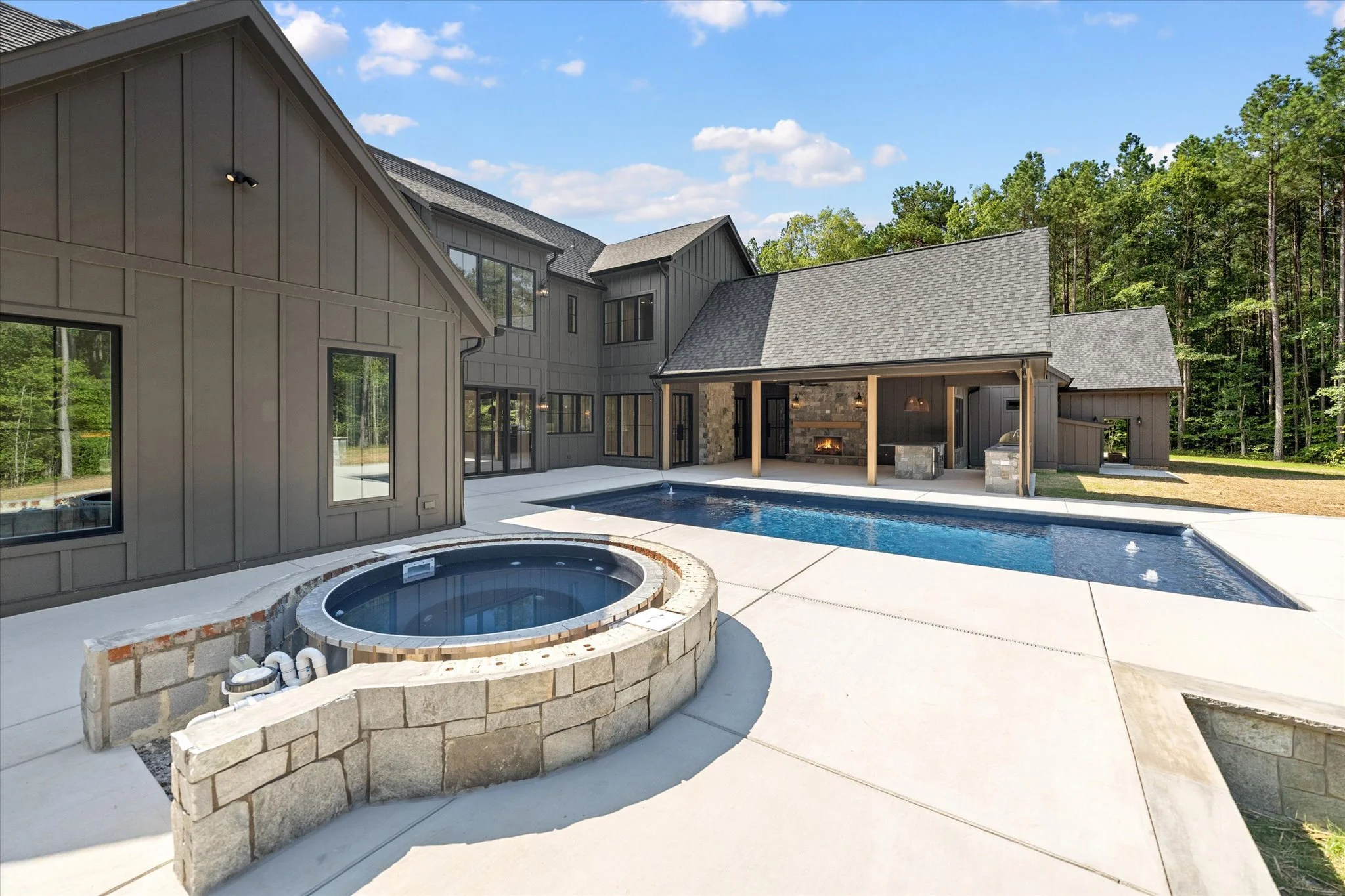 Modern house with a backyard featuring a swimming pool, hot tub, fire pit, and a large patio area, surrounded by a wooded area under a blue sky with clouds.