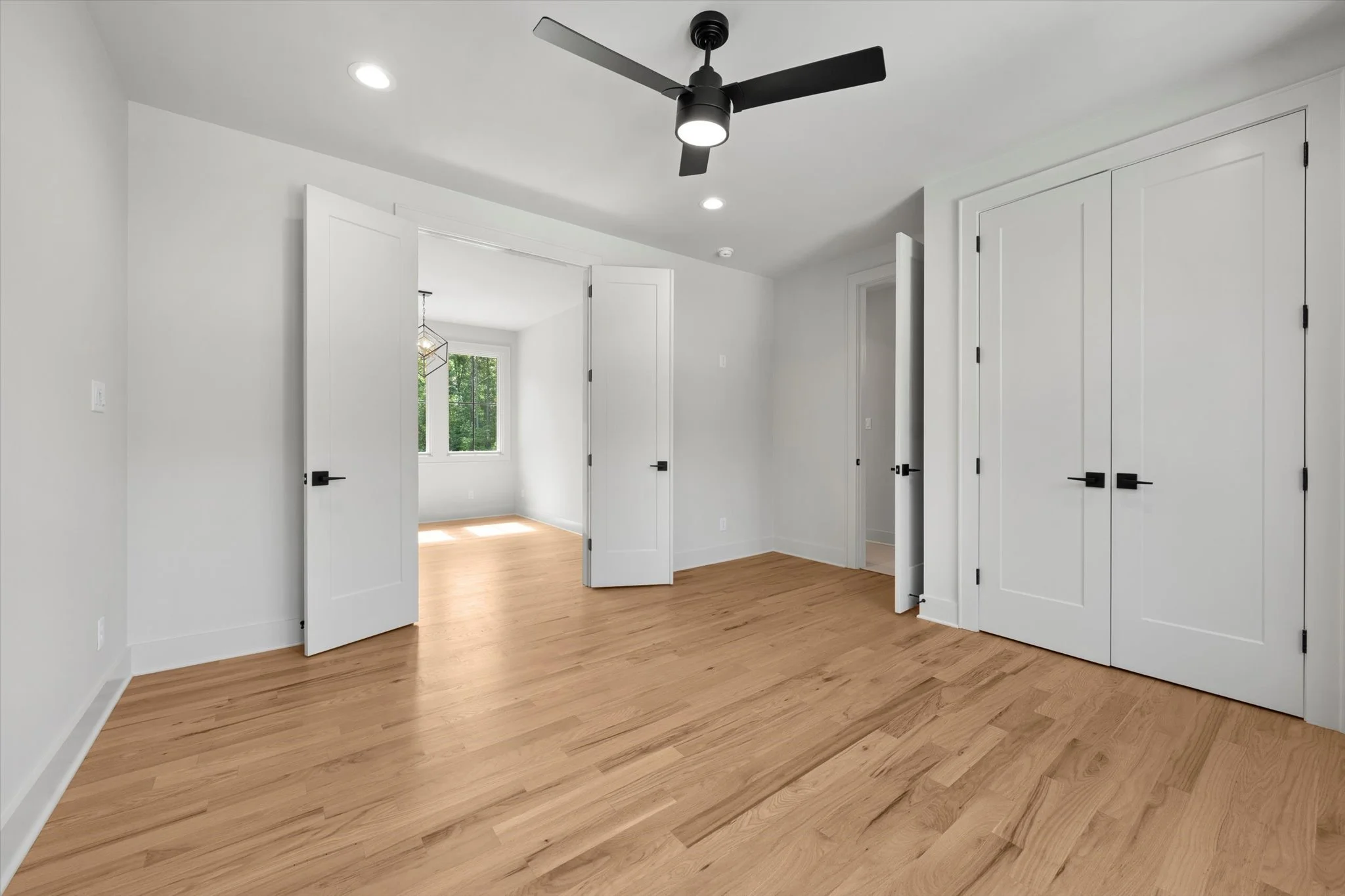 Empty bedroom with white walls, hardwood floor, ceiling fan, and closet doors, with an adjoining room visible through open doors.