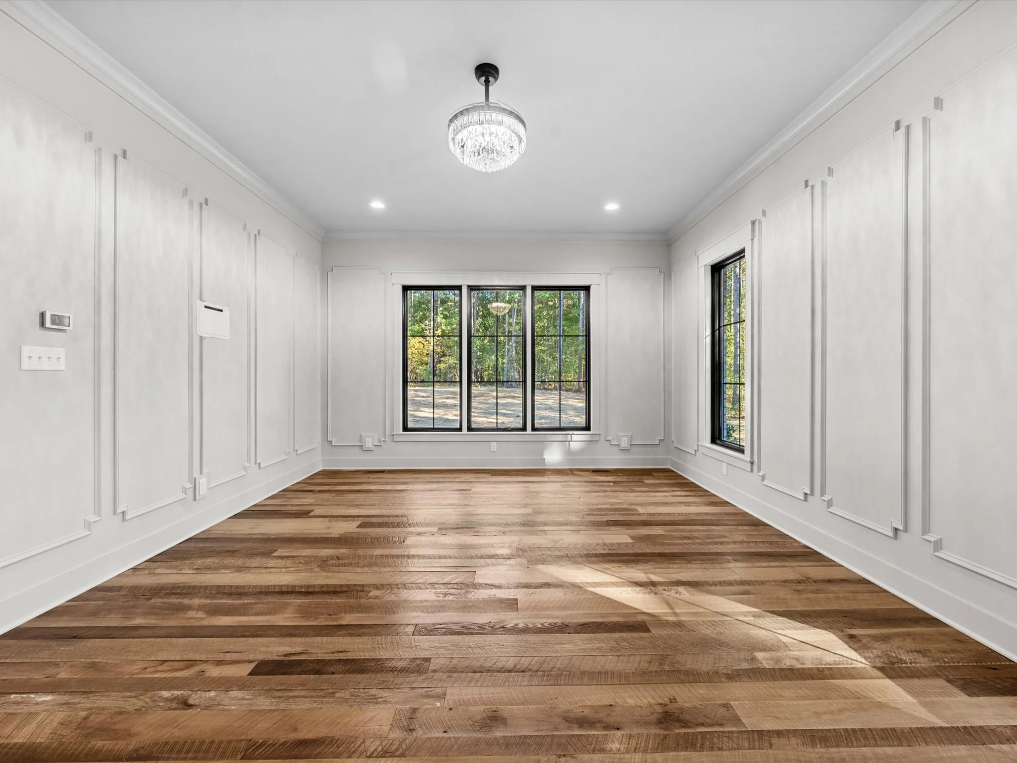 Empty room with hardwood floors, white walls, large window, and ceiling chandelier.