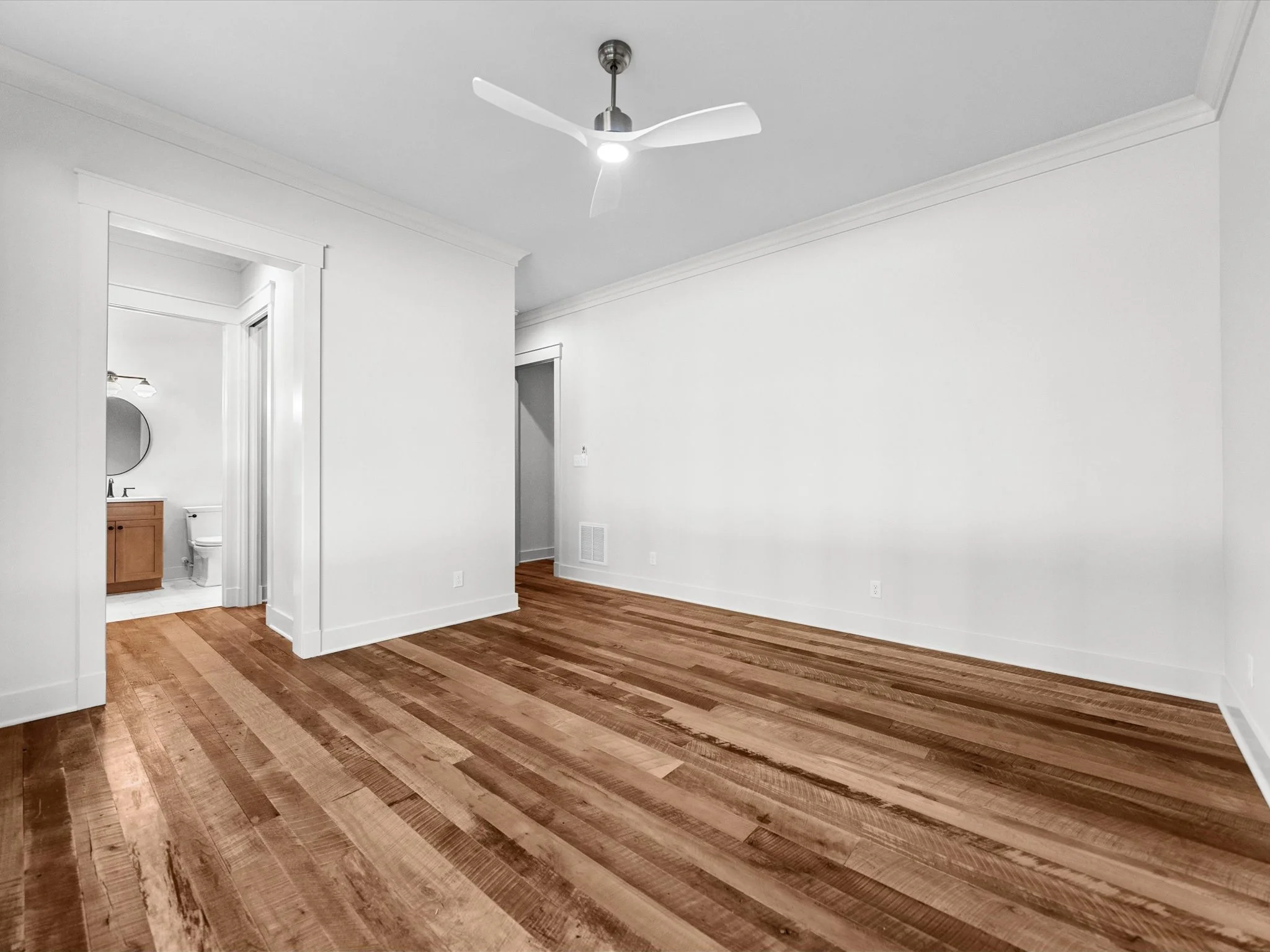 Empty living room with white walls, wood flooring, ceiling fan, and adjoining bathroom with wooden vanity and mirror.