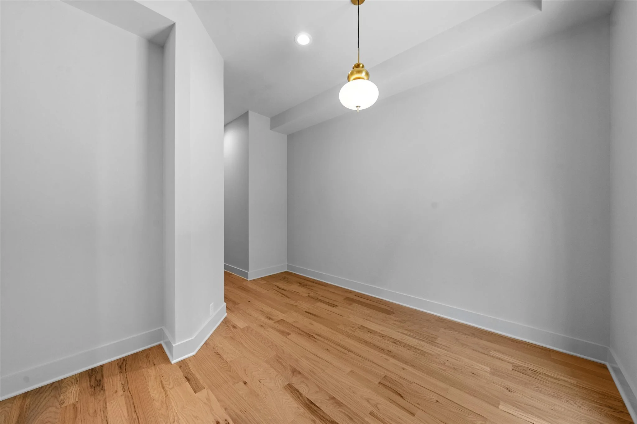 Empty room with white walls, hardwood flooring, and a hanging light fixture with a spherical shade.