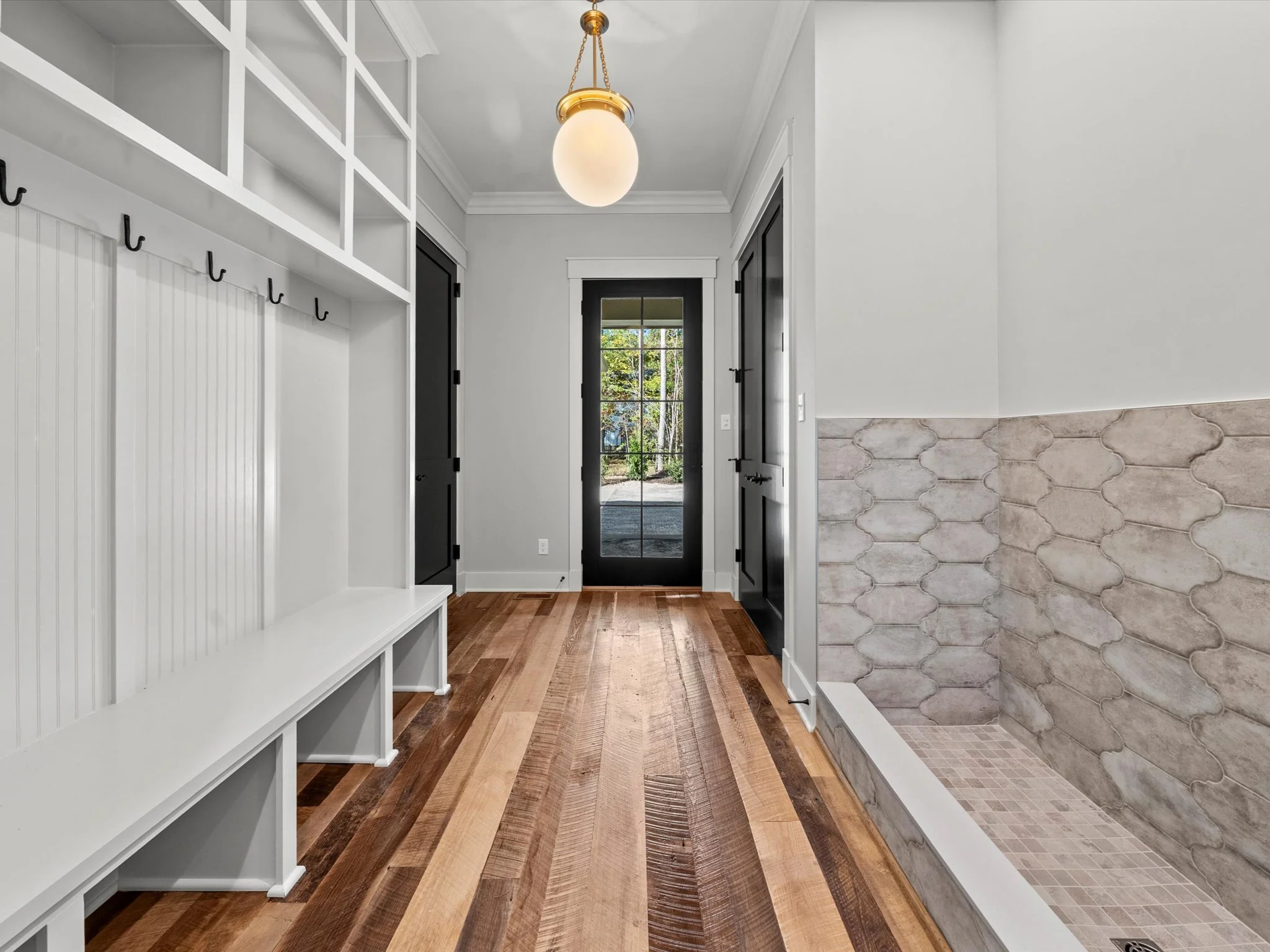 Entryway with hardwood flooring, black door with glass panes, white walls, and a built-in white bench and coat hooks on the left. To the right, a tiled section with neutral-colored tiles, likely a shower or utility area.