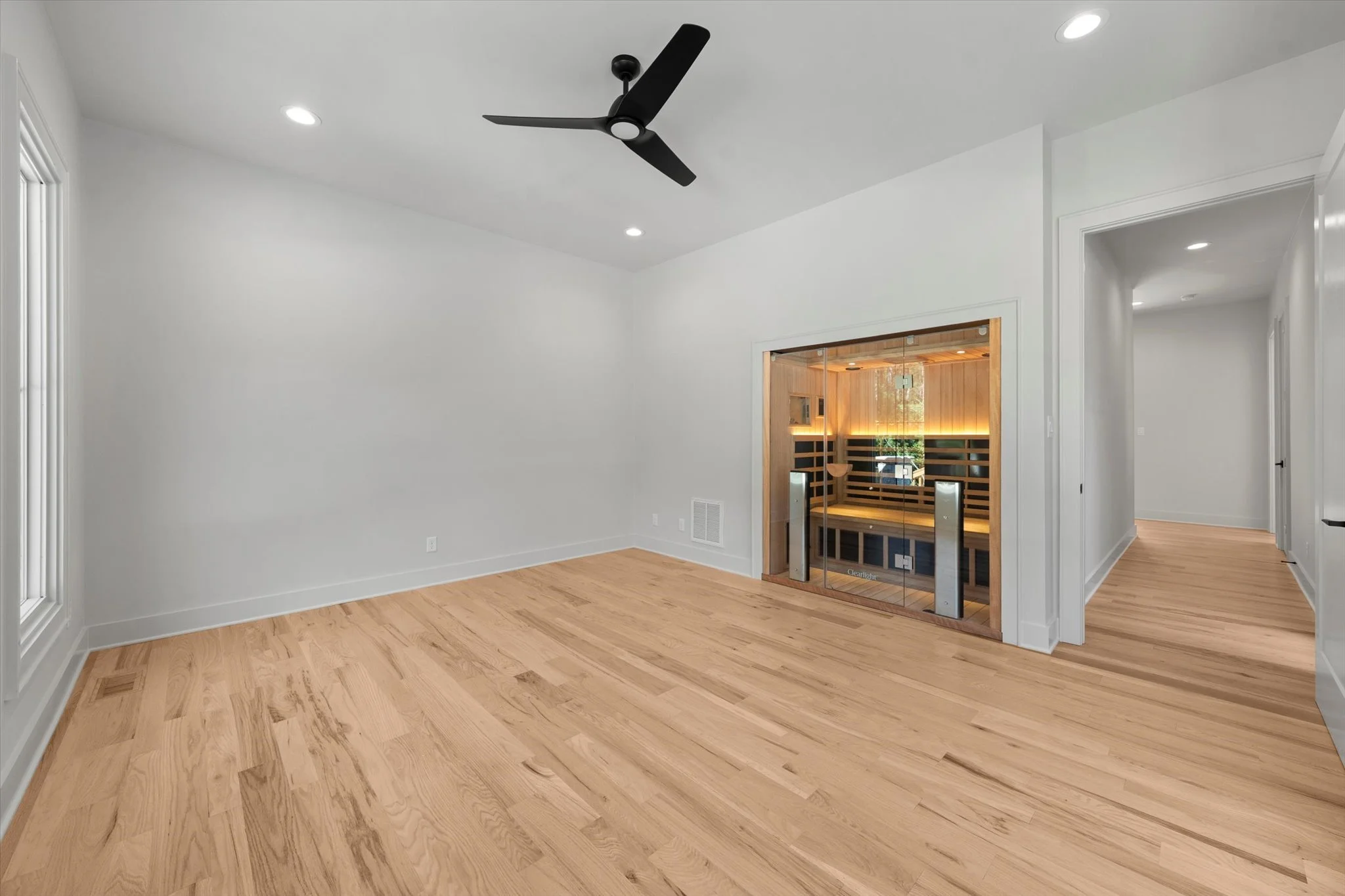 Empty room with light wood flooring, white walls, recessed lighting, a ceiling fan, and a doorway leading to a sauna with glass door and wooden interior.