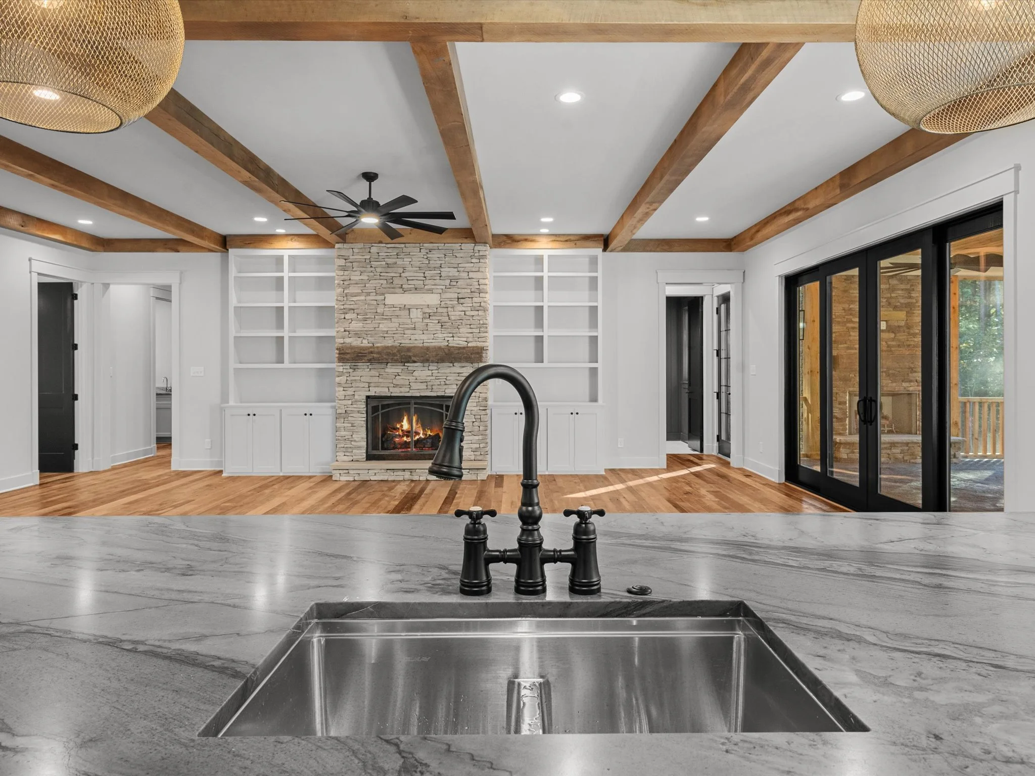 Modern living room with a stone fireplace, wooden beams on ceiling, white built-in shelves, hardwood floor, black ceiling fan, sliding doors to outdoors, kitchen sink with black faucet in foreground.