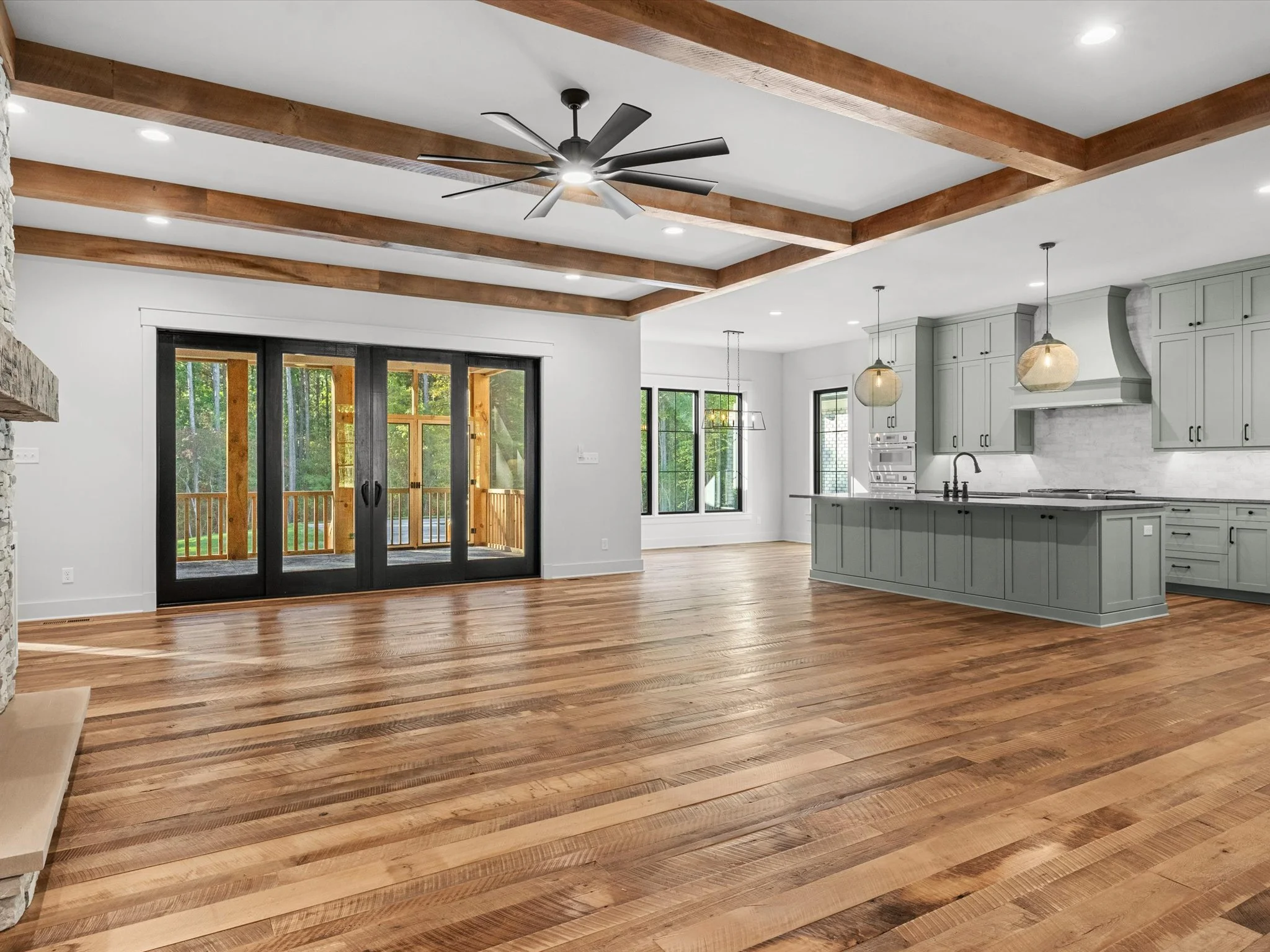 Open-concept living room and kitchen with hardwood floors, gray cabinetry, large sliding glass doors leading to a deck, and wooden beams on the ceiling.