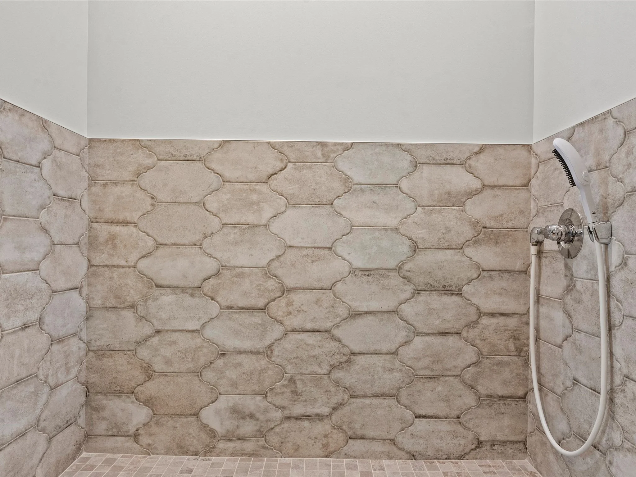 A shower area with beige patterned tile walls, a handheld showerhead, and a white ceiling.