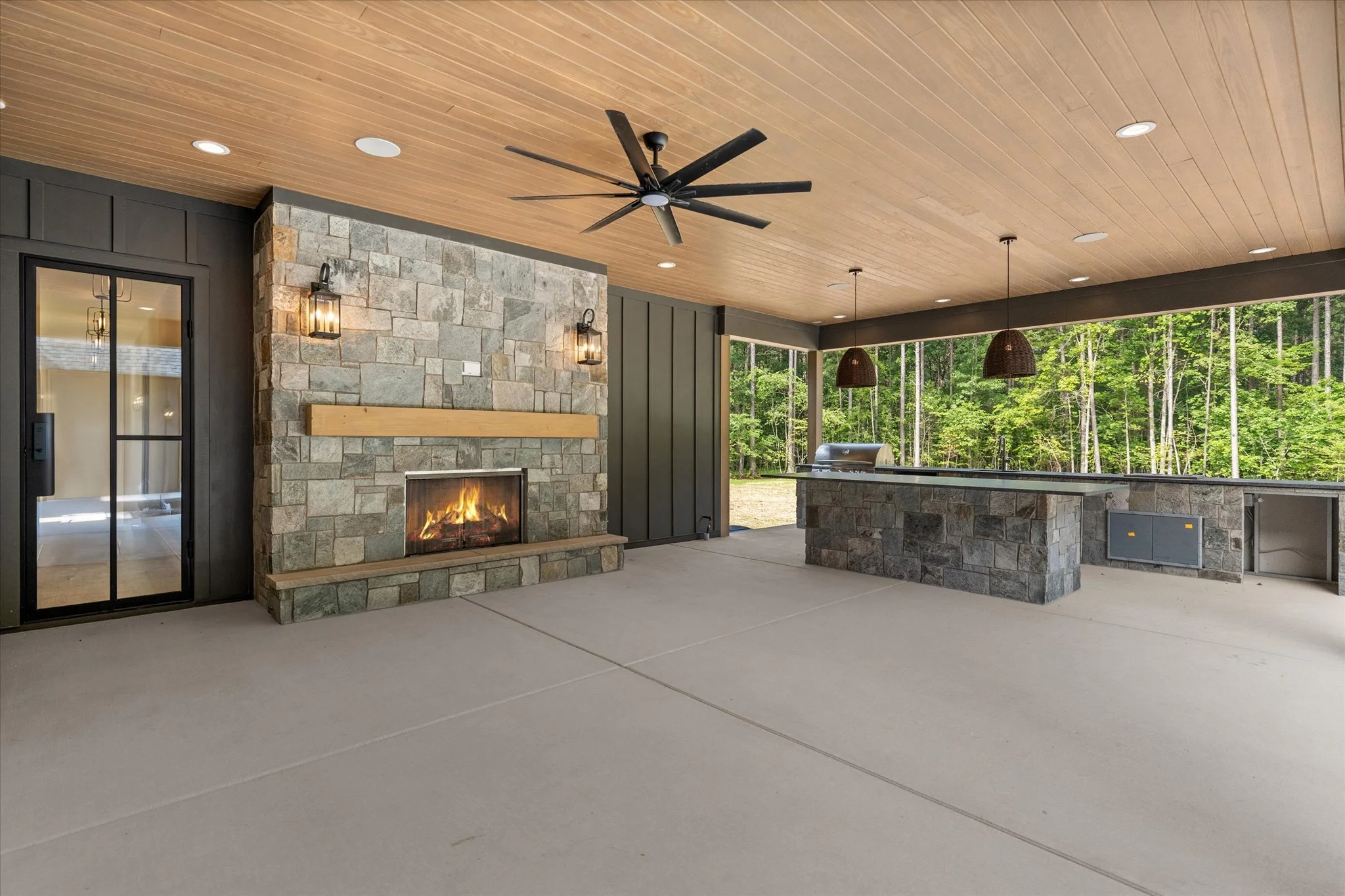 Outdoor patio with stone fireplace, ceiling fan, and kitchen area, overlooking a wooded backyard.