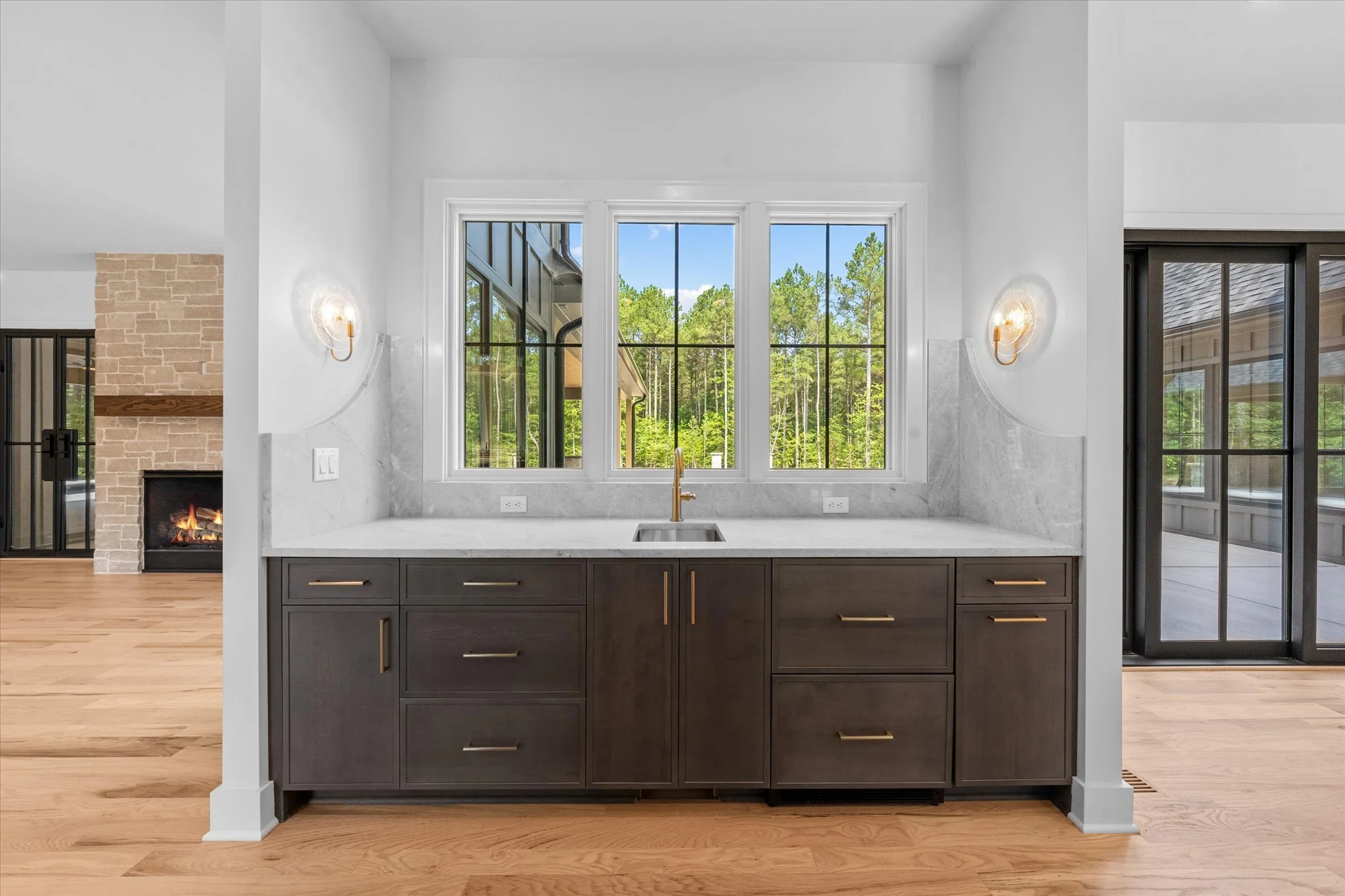 Modern kitchen with dark wood cabinets, white marble countertop, gold fixtures, and a large window overlooking outdoor greenery.