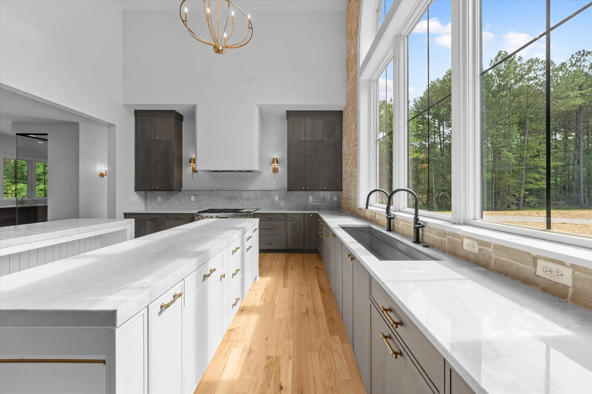 Modern kitchen with large window, gray cabinets, white countertops, and brass hardware, overlooking a wooded outdoor area.