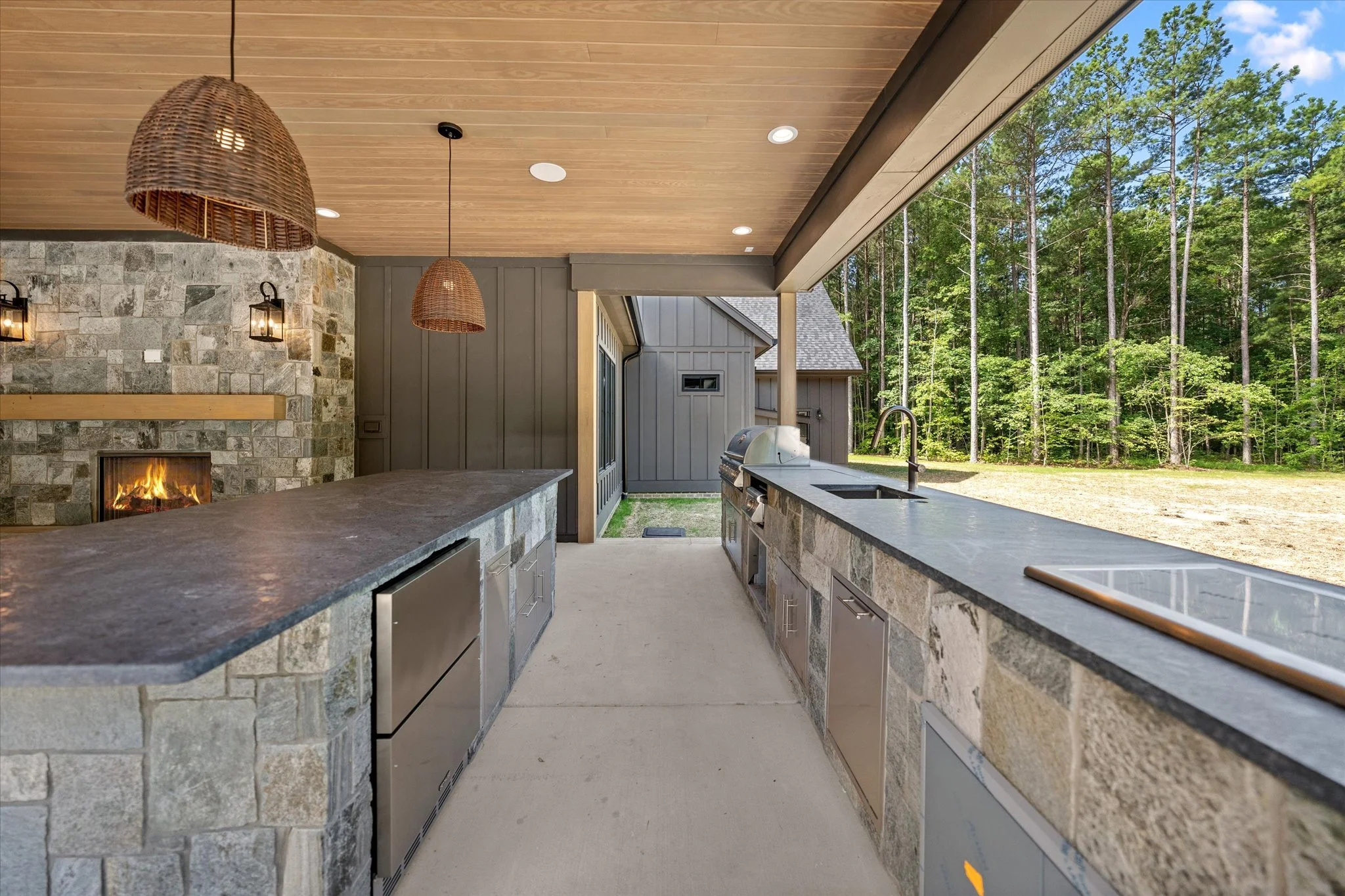 Outdoor kitchen with stone countertops, stainless steel appliances, a sink, and a barbecue grill, overlooking a forested yard.