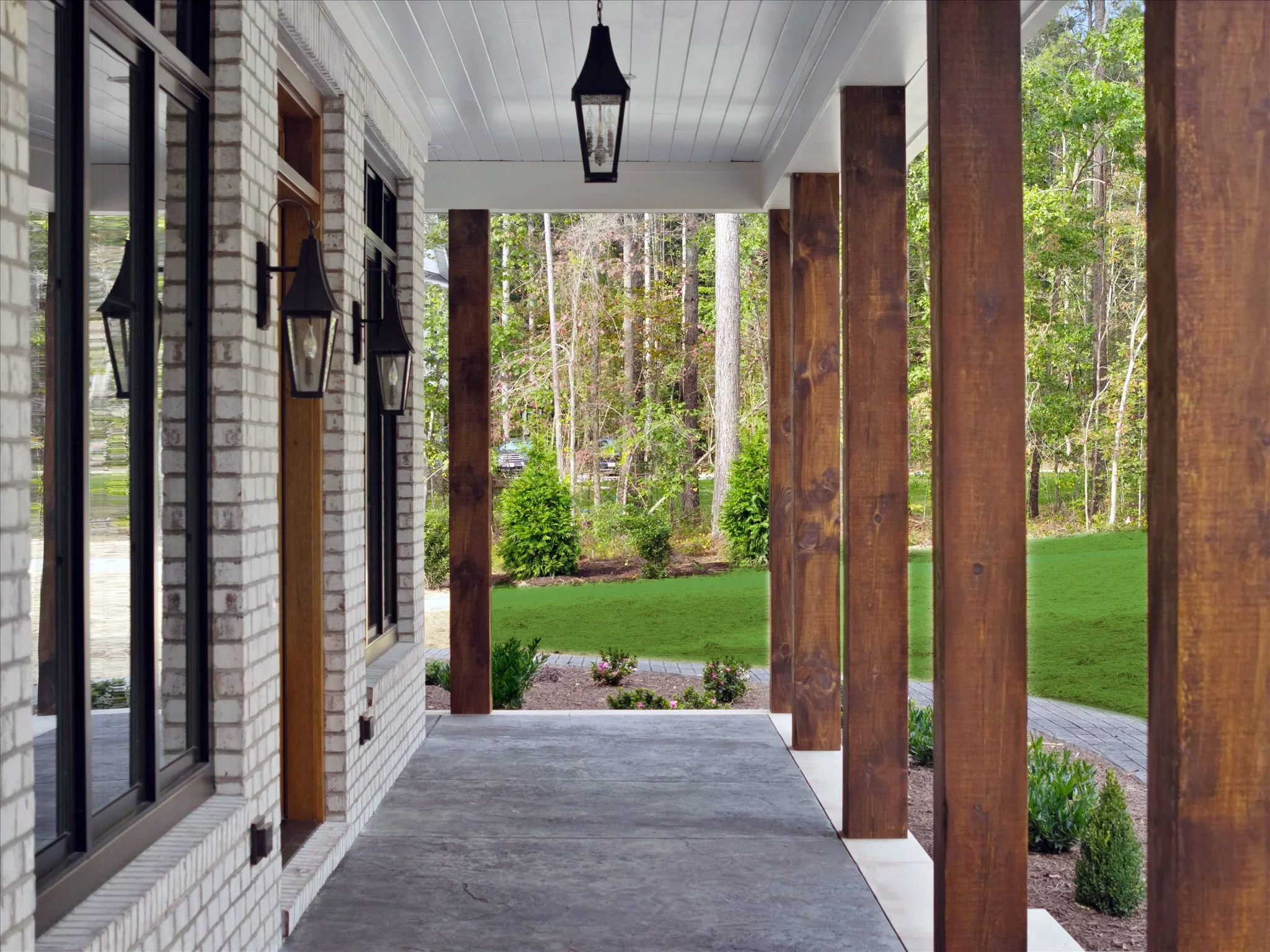 View of a porch with wooden posts, brick wall, large windows, hanging lanterns, and a landscaped yard with green grass and trees in the background.