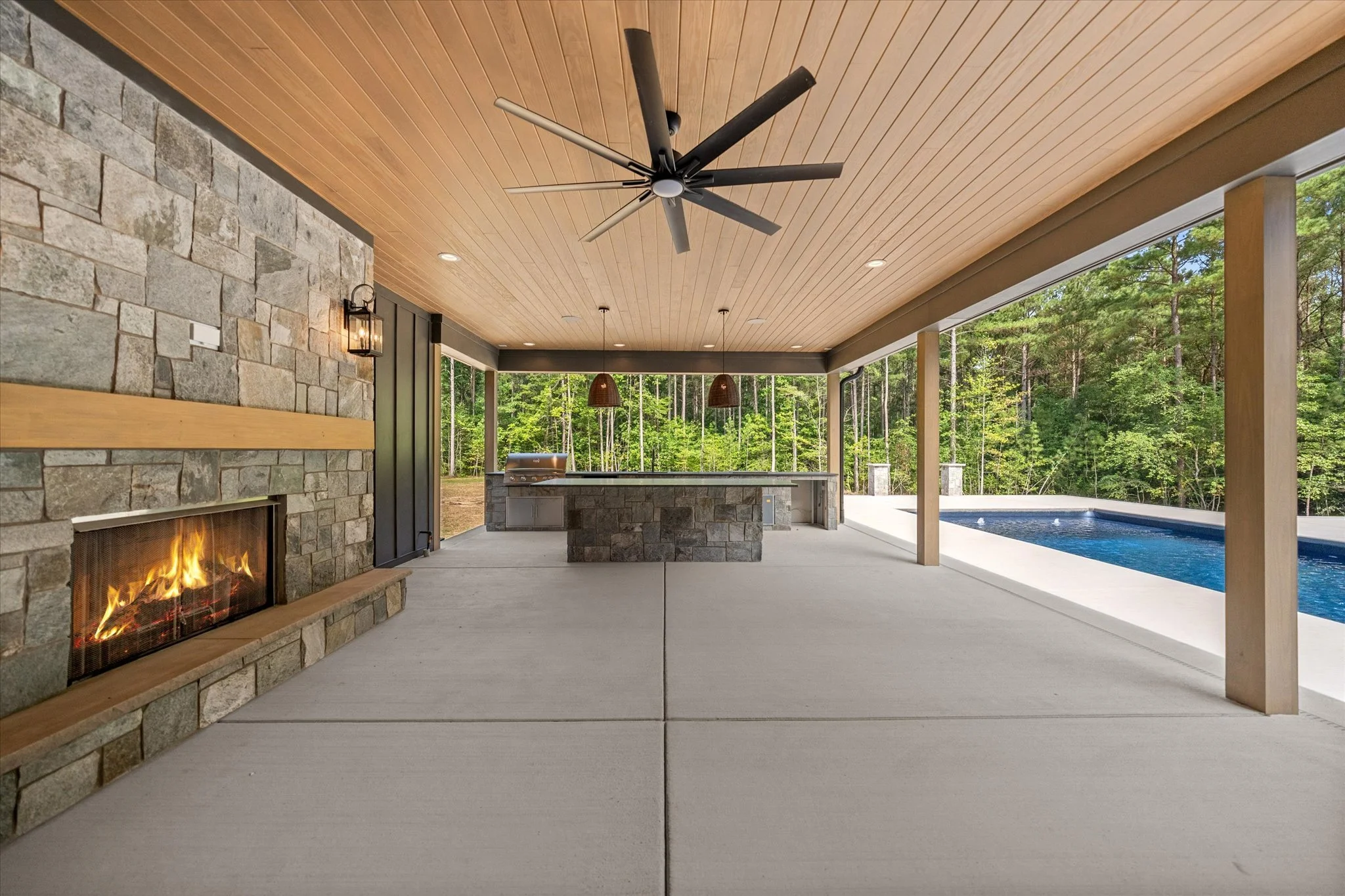 Covered patio with stone fireplace, ceiling fan, outdoor kitchen, and swimming pool surrounded by trees.