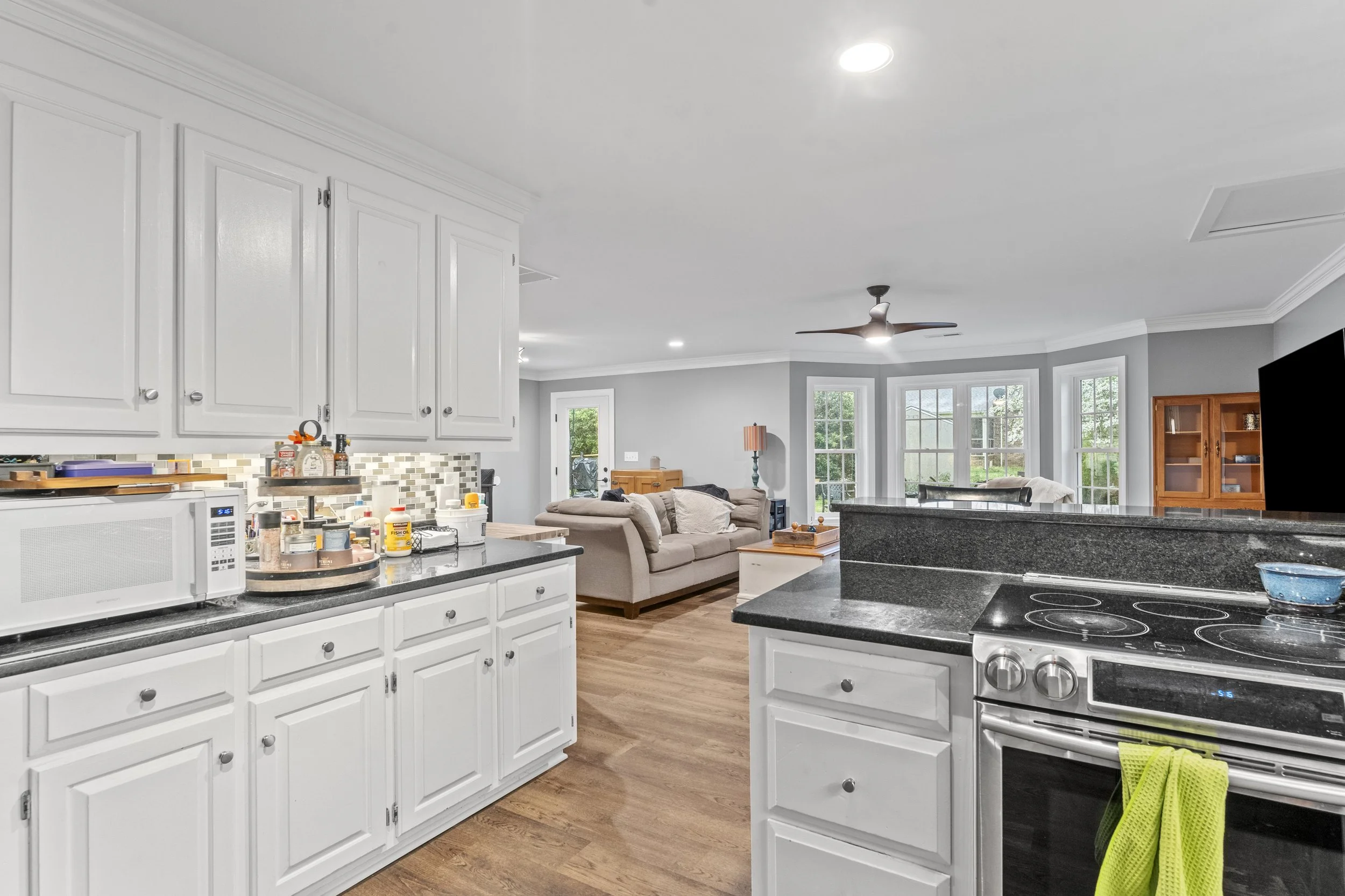 Open kitchen and living room with white cabinets, black countertops, wood flooring, beige sofa, and large bay windows with a view of the backyard.
