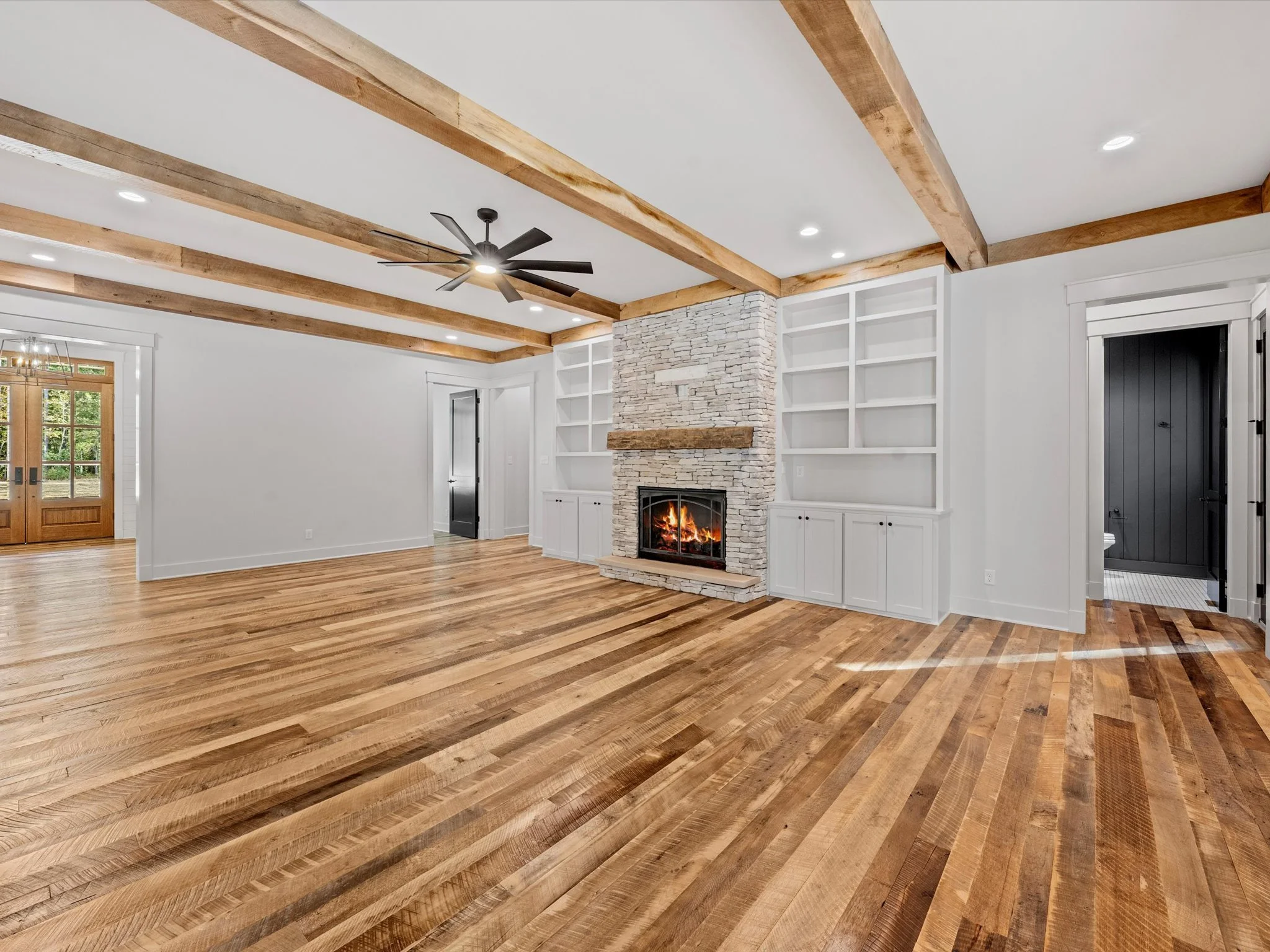 Living room with white walls, wooden beams on the ceiling, hardwood floor, and a stone fireplace with a fire burning. Built-in white shelves on each side of the fireplace. Door to the outside and a doorway leading to a bathroom or closet visible.