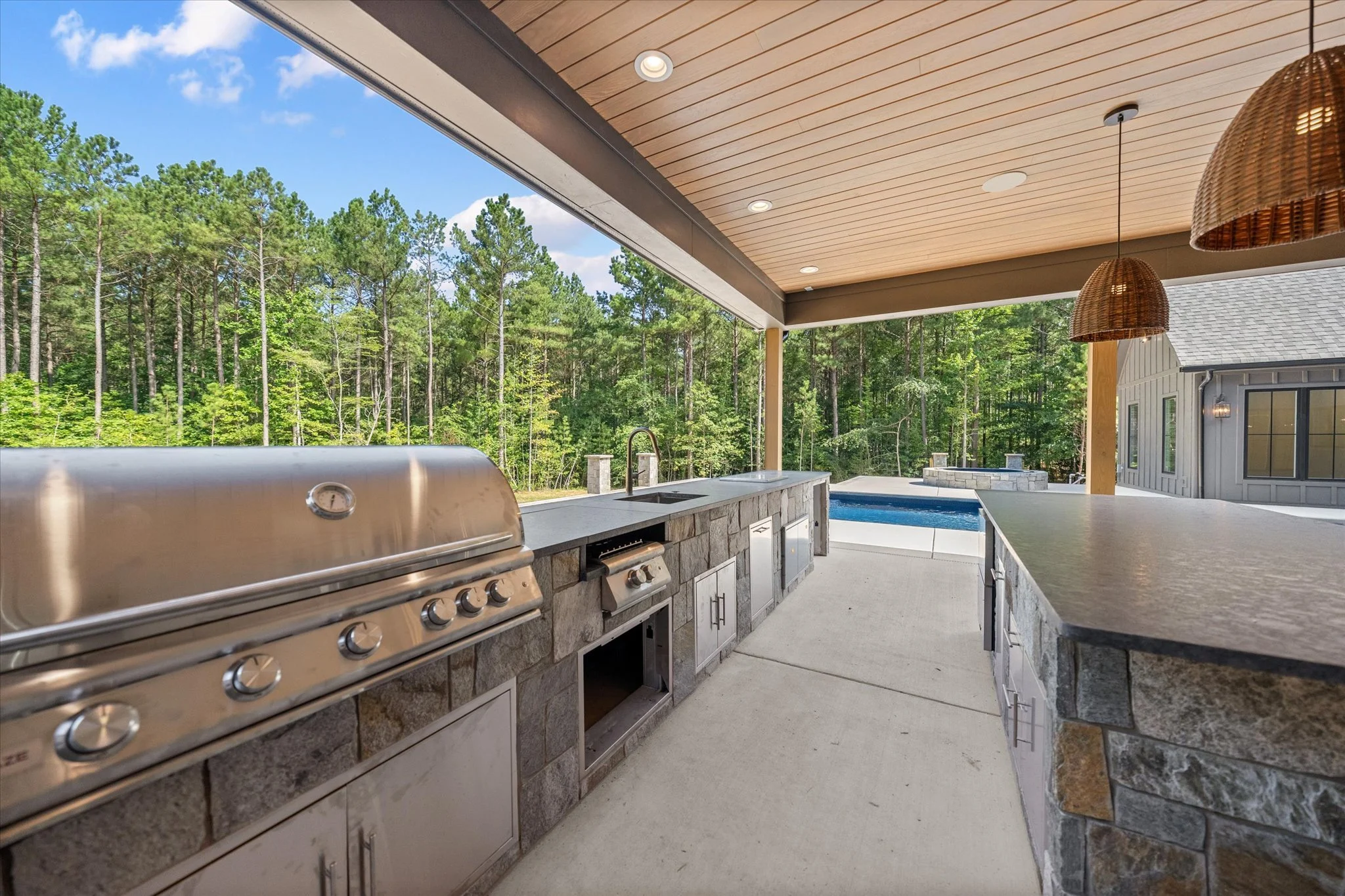 Outdoor kitchen with a built-in grill, sink, and stone countertops, overlooking a backyard with a pool and surrounded by trees.