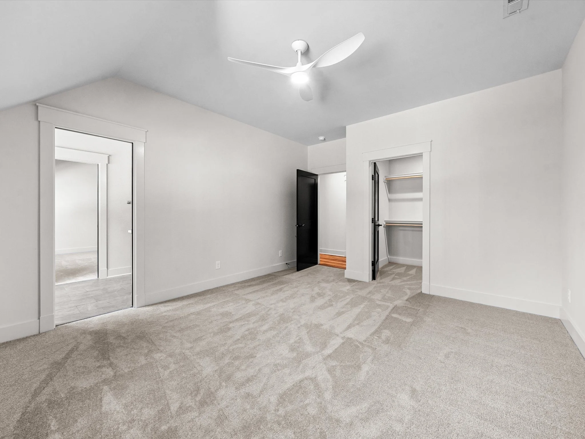 Empty bedroom with beige carpet, white walls, ceiling fan, open closet with wire shelving, and door leading to hall with wood flooring.