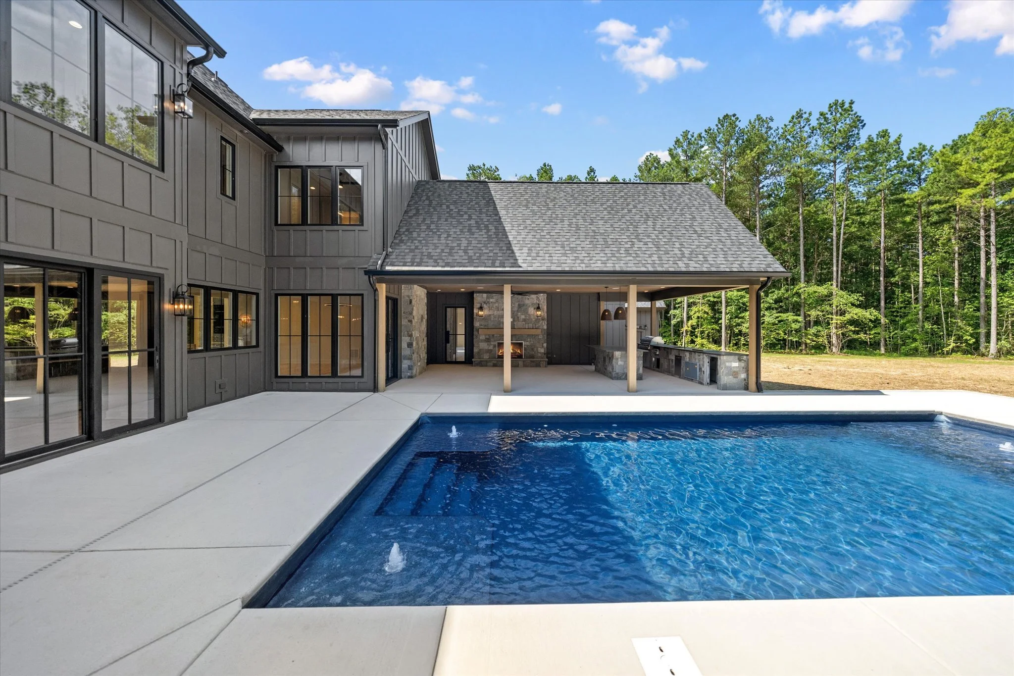 Modern house with black exterior, large windows, outdoor fireplace, and a swimming pool in a backyard surrounded by trees.