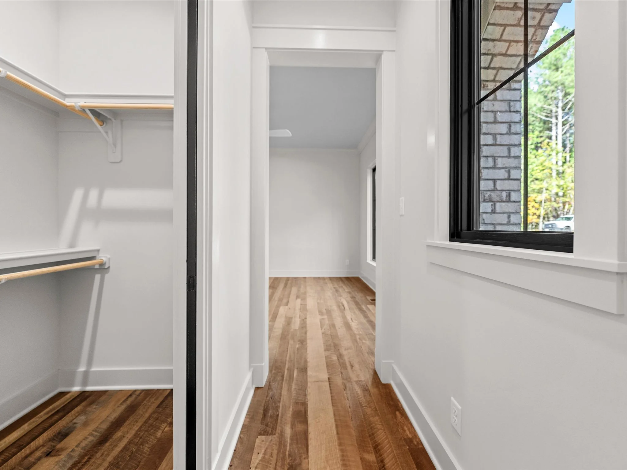 Empty walk-in closet with white walls, wooden flooring, and a window with a black frame showing trees outside.