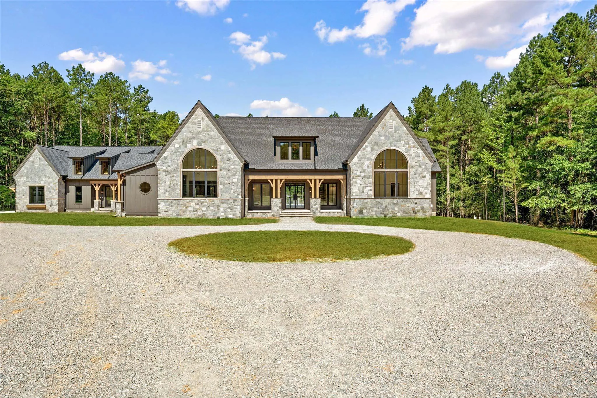 Large stone house with multiple gables, large arched windows, and a gravel driveway, surrounded by green trees under a partly cloudy sky.