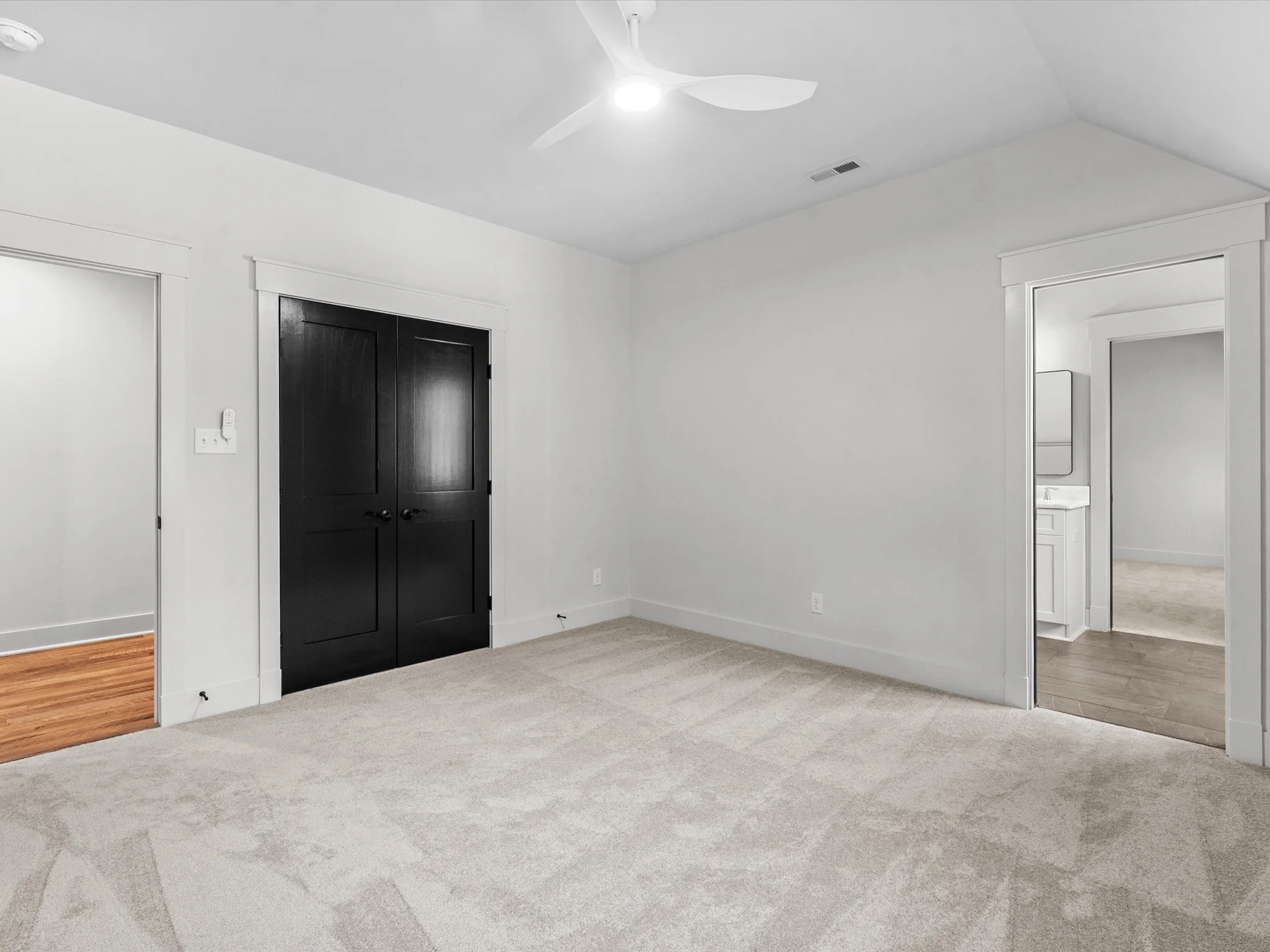 Empty bedroom with white walls, beige carpet, black closet doors, and an adjacent bathroom with a white vanity and mirror.