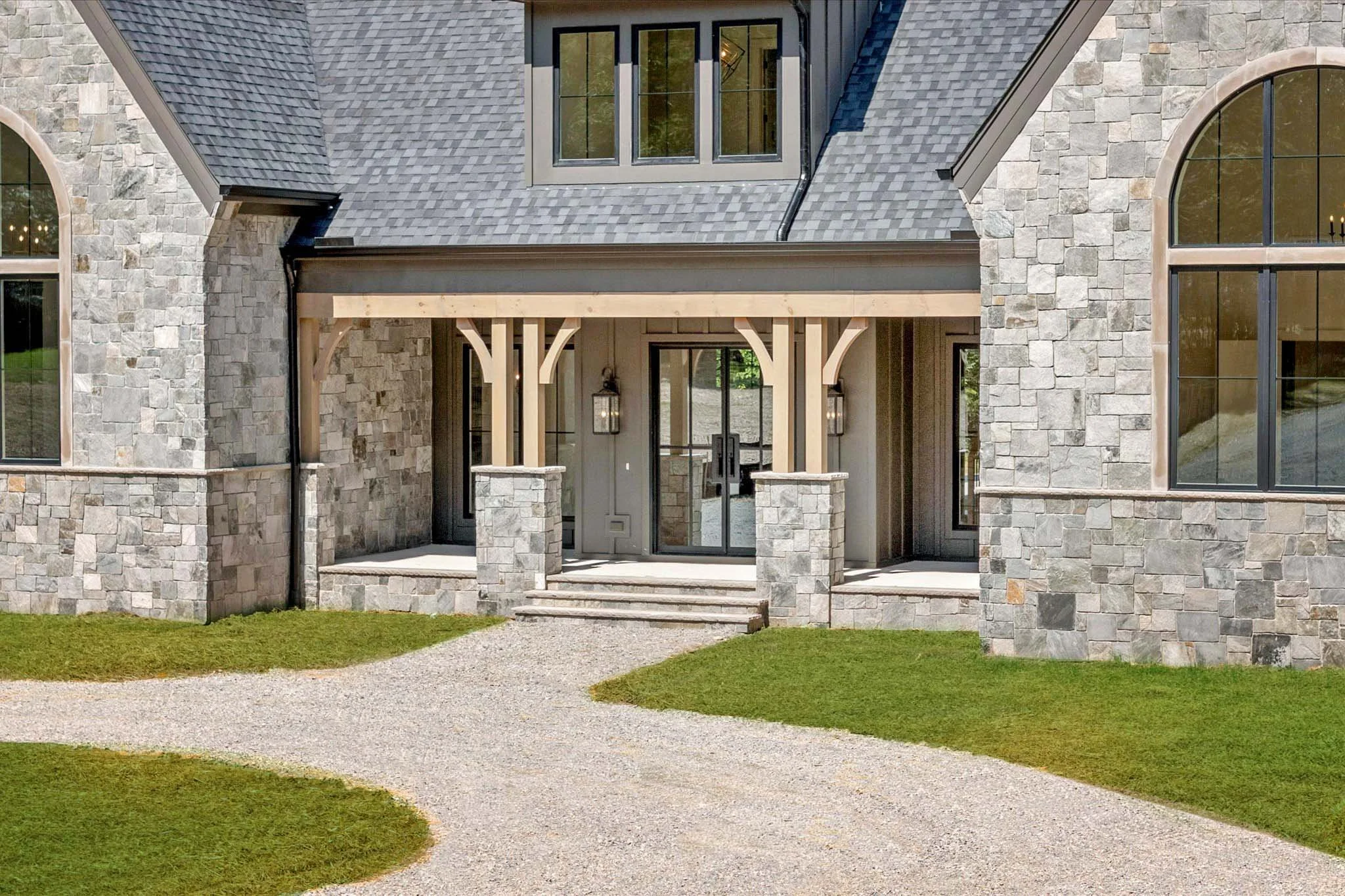 Front entrance of a stone house with a porch supported by wooden pillars, set on a gravel pathway with grass on either side.