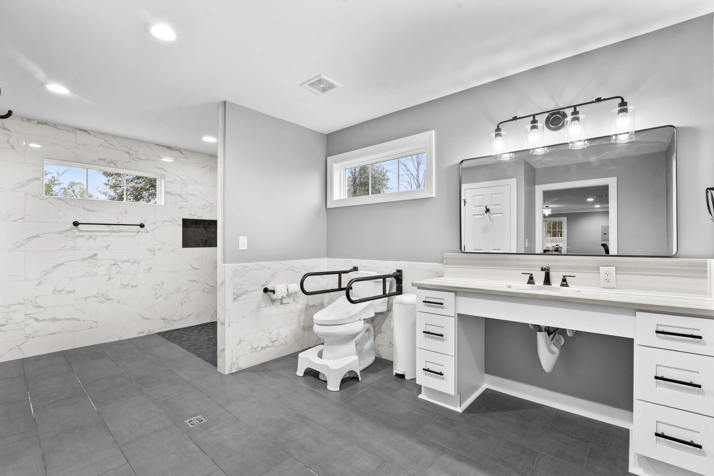 Bathroom with a walk-in shower, wheelchair accessibility features, a toilet, a vanity with a large mirror, and a window showing trees outside.