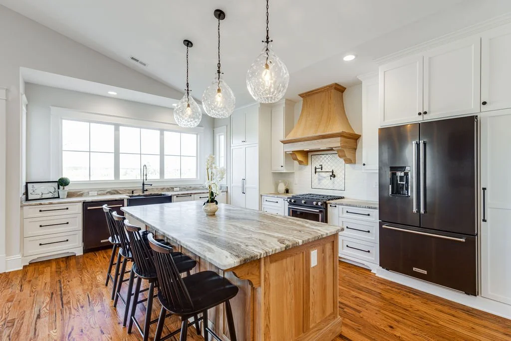 Modern kitchen with white cabinets, a large island with a granite countertop, black bar stools, stainless steel refrigerator, black oven with a wood range hood, and pendant lights. Large window above the sink.