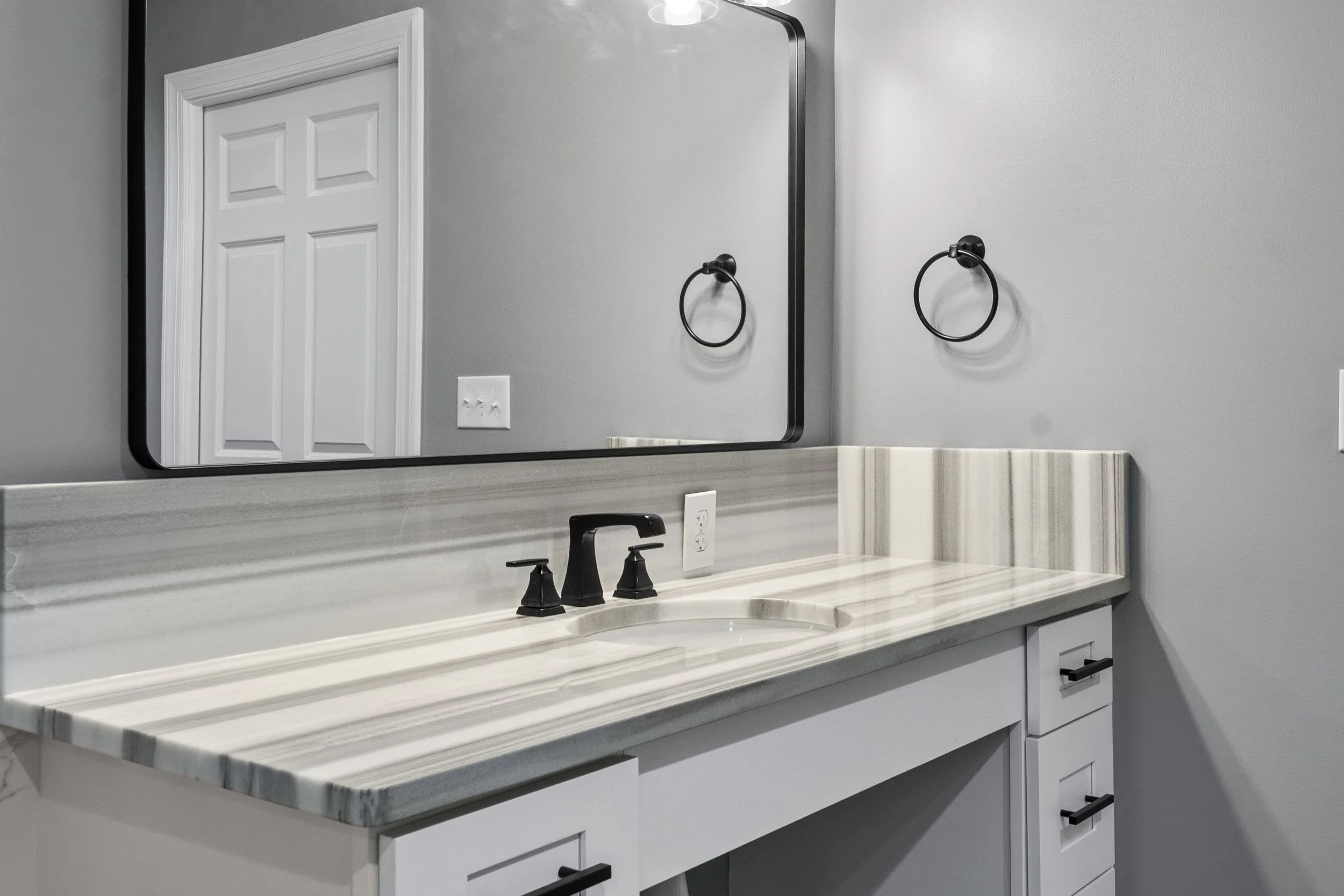 Bathroom vanity with a marble countertop, a black faucet, a large rectangular mirror, and black towel rings on a gray wall.
