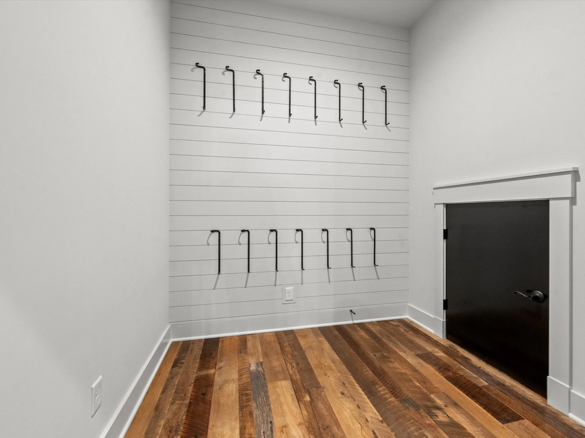 Empty room with wooden floor, white walls, a wall with hooks, and a black door.