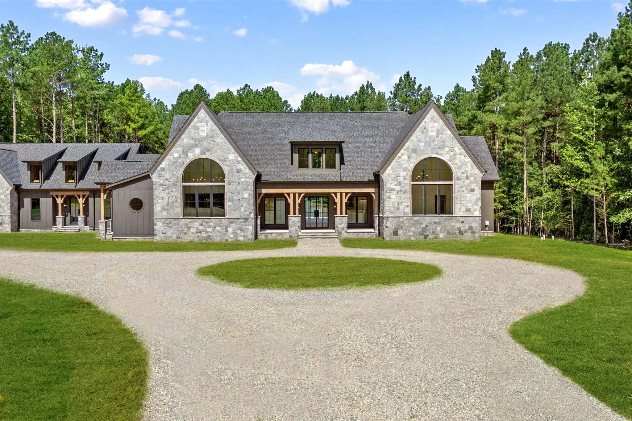 A large stone and wood house surrounded by a circular gravel driveway and lush green grass, with a wooded area in the background.