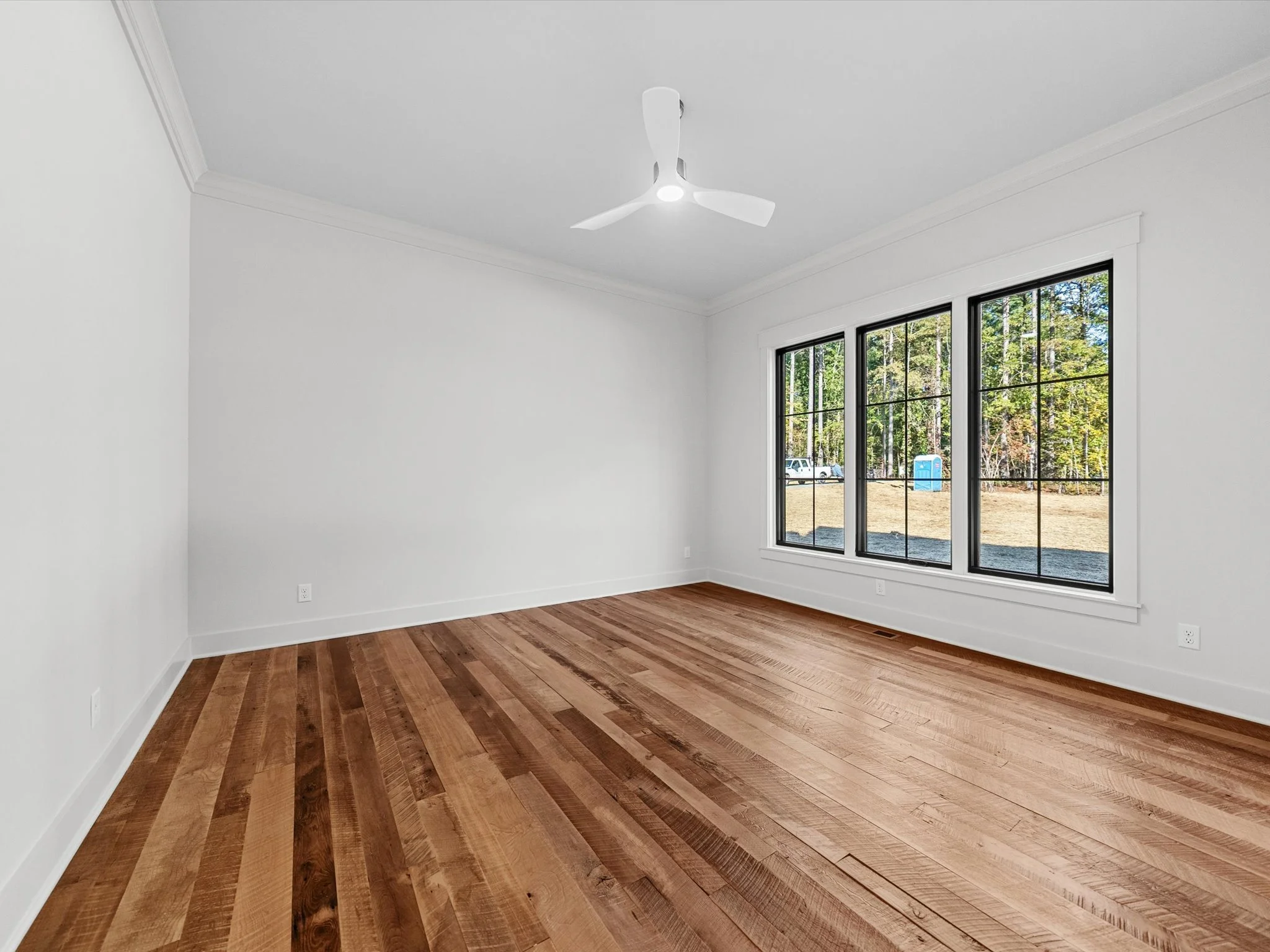 Empty room with white walls, wooden floor, large windows, a ceiling fan, and a view of trees outside.