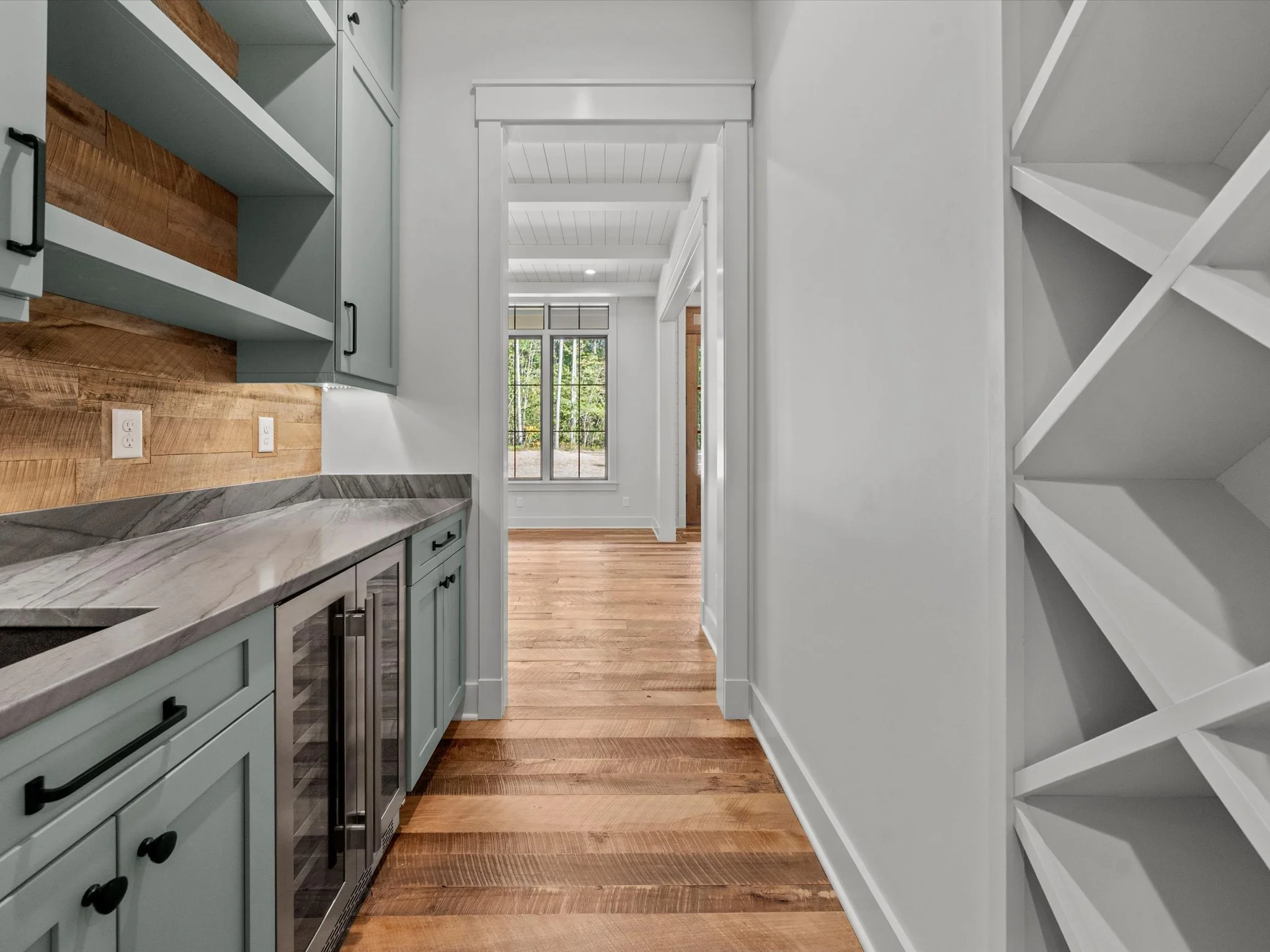 Interior view of a kitchen with teal cabinets, a marble countertop, a wooden backsplash, and built-in wine cooler, leading to a bright room with wooden floors and large windows.