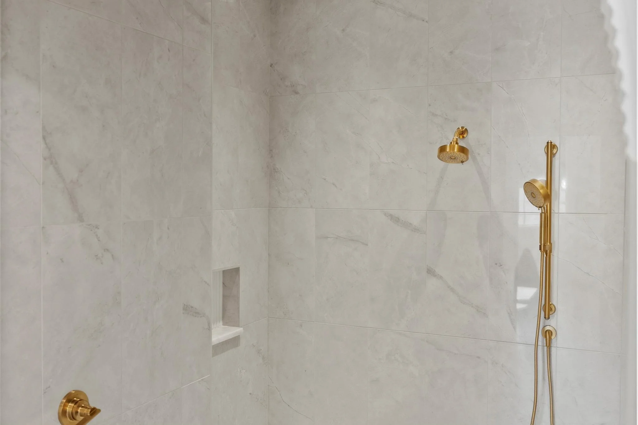 A minimalist bathroom shower with large white marble tiles, a built-in niche, and gold-colored shower fixtures including a handheld showerhead and a fixed showerhead.