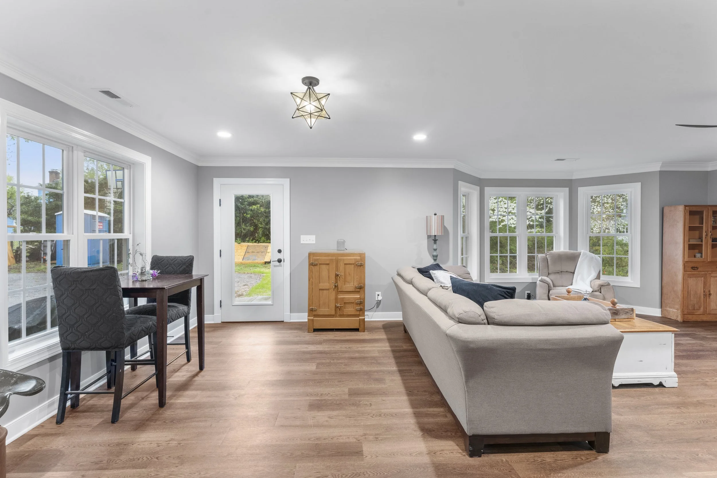 Living room with beige sofa, gray walls, hardwood floors, large windows, small dining table with two chairs, wooden cabinet, lamp, and door leading outside.