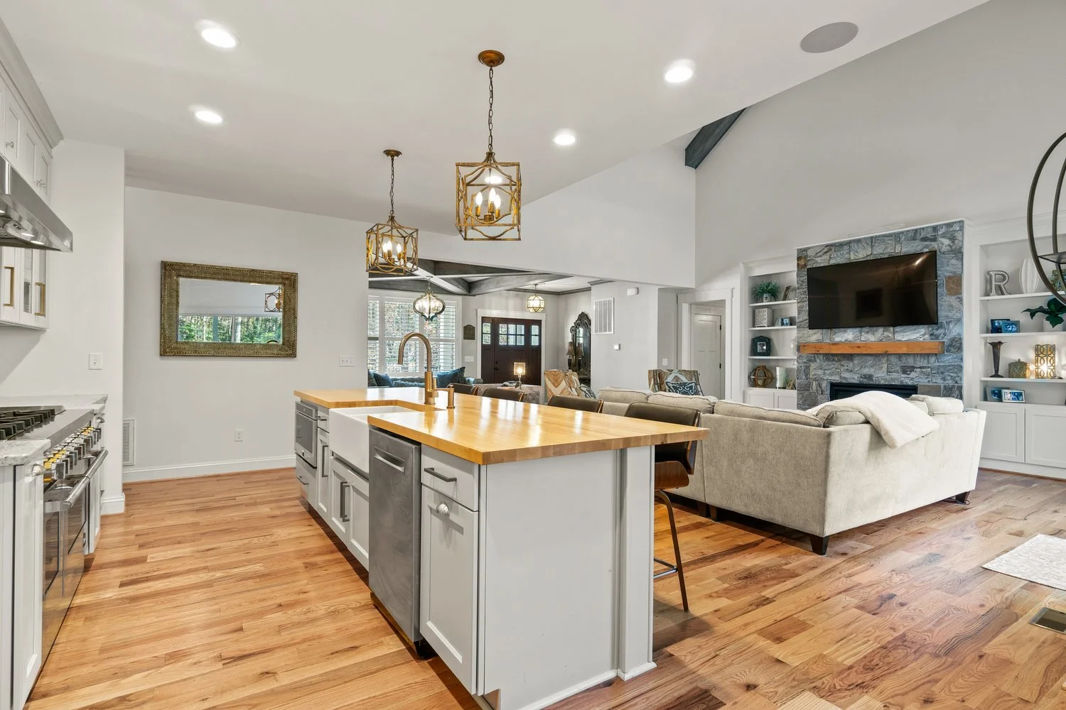 Open-concept living space with kitchen and living room, wooden flooring, white cabinets, kitchen island with wooden countertop, gray upholstered sofa, stone fireplace with TV, built-in shelves with decorative items, and large windows.
