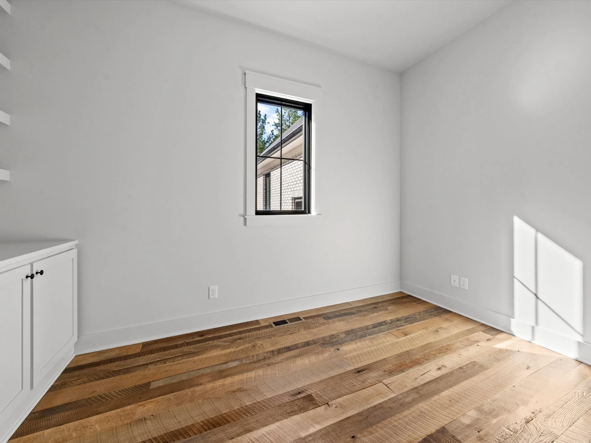 Empty room with white walls, wooden floor, window showing an exterior brick building, and built-in white cabinet on the left.
