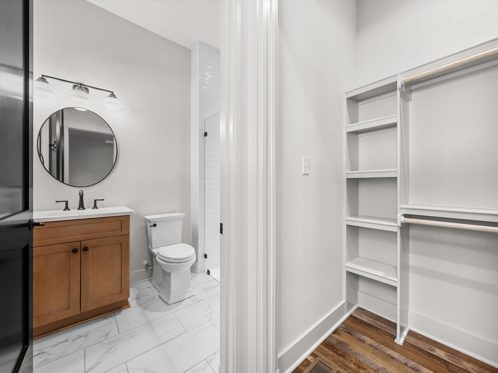 Bathroom with white walls, a wooden vanity with a white countertop, an oval mirror, a light fixture with three bulbs, a white toilet, a walk-in shower with white tiles, and an open white closet with shelves and a hanging rod.
