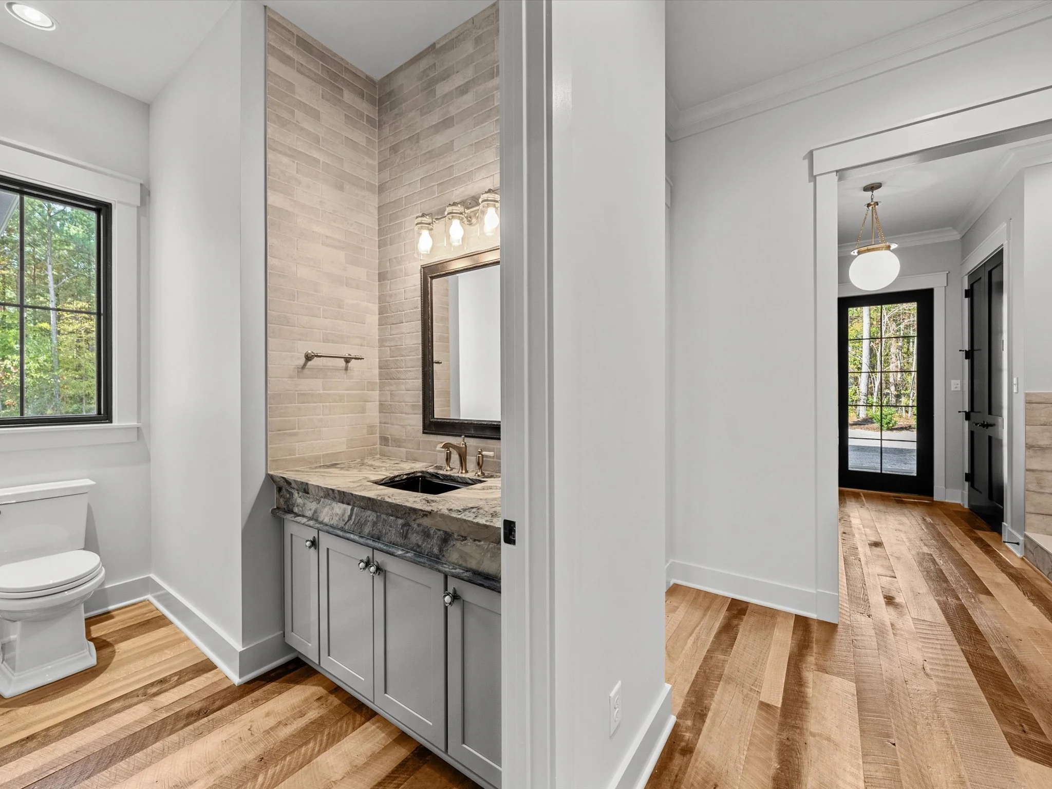 A modern bathroom with a toilet, a vanity with a granite countertop, a mirror, and a light fixture. The room has a window and wood flooring. Adjacent is an entryway with a decorative light fixture and glass door, featuring a black door frame and wood