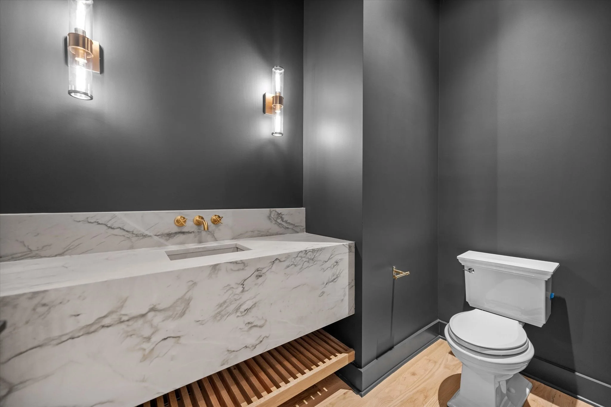 Modern bathroom with dark gray walls, white marble sink with gold fixtures, and white toilet on a wooden floor.