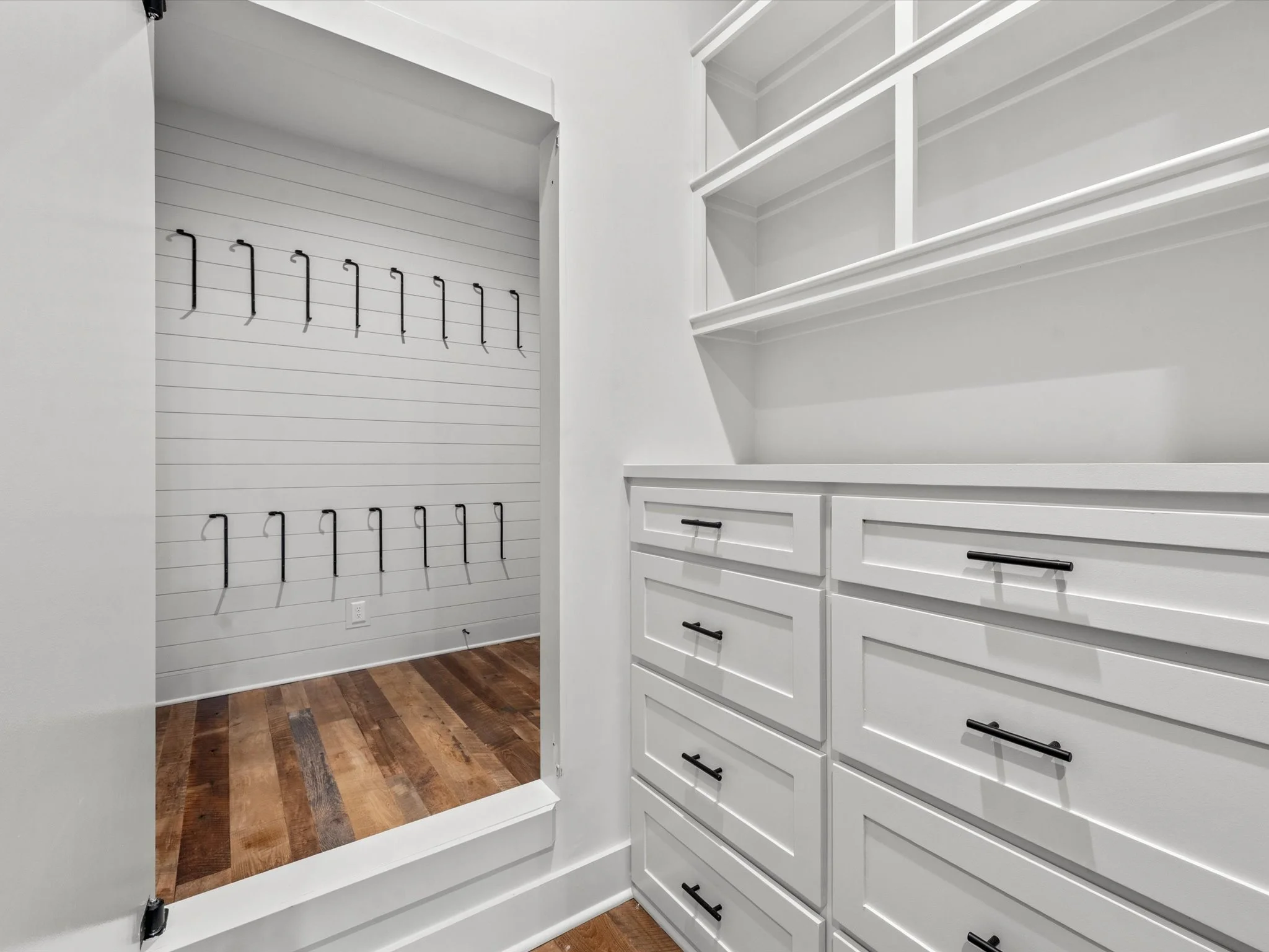 A white walk-in closet with built-in drawers, open shelving, and black handles; an adjacent small room with black hooks on the wall, a white shiplap accent wall, and hardwood flooring.