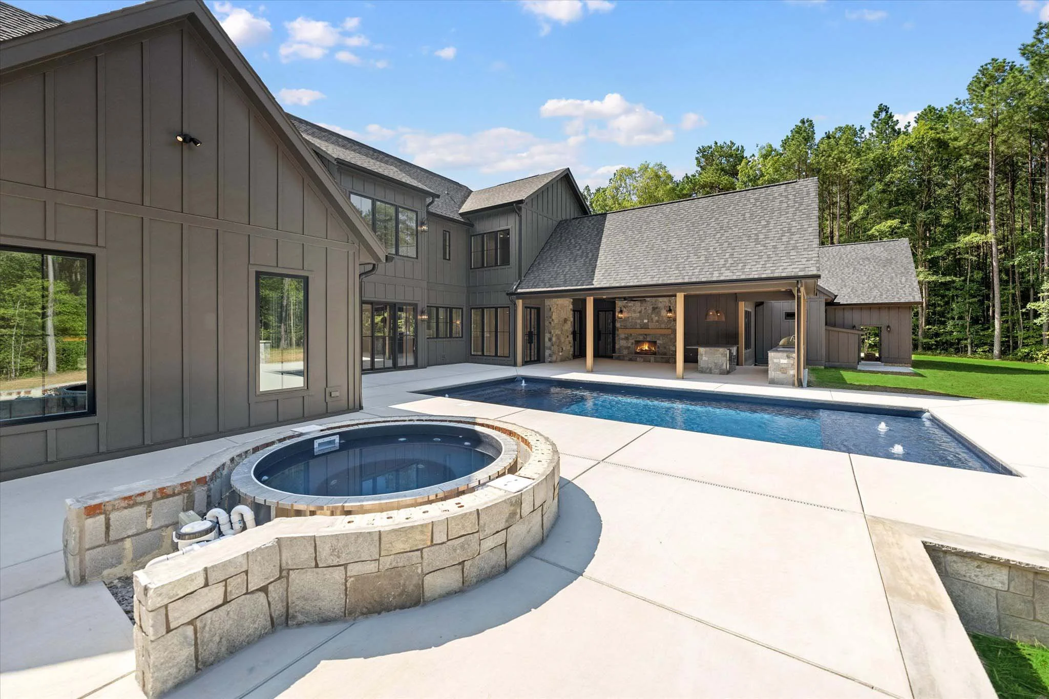 Modern backyard with a swimming pool, hot tub, and an outdoor fireplace under a covered patio, with a large house and wooded background.