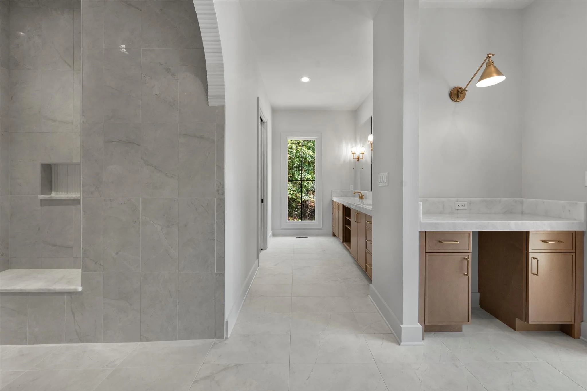 A modern bathroom with a glass-enclosed shower on the left, white walls, light-colored tile flooring, a wooden vanity with a white countertop and gold fixtures, a wall-mounted gold lamp, and a window showing green foliage outside.